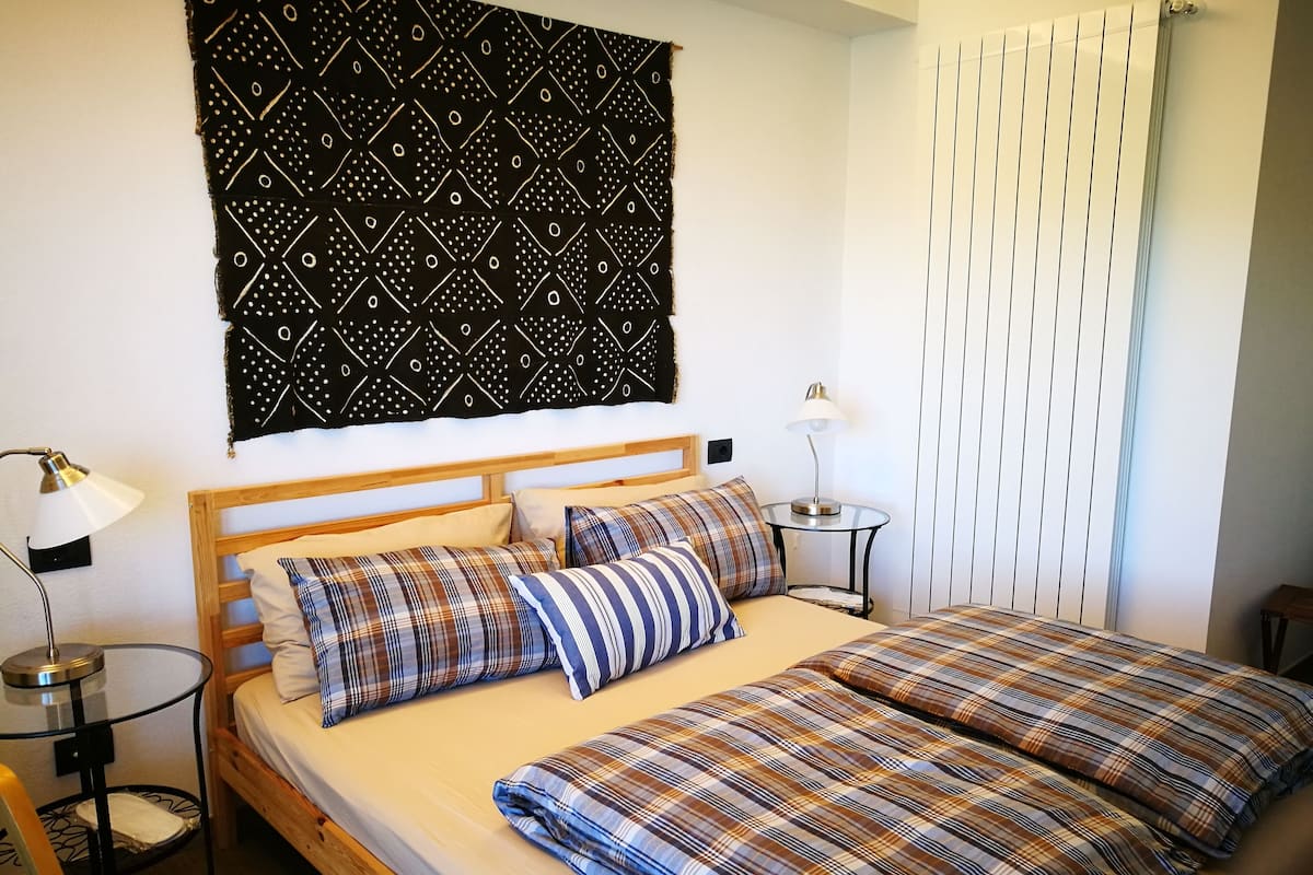 Una cama doble con ropa de cama escocesa, una mesita de noche con lámpara, un tapiz estampado y una ventana con persianas.