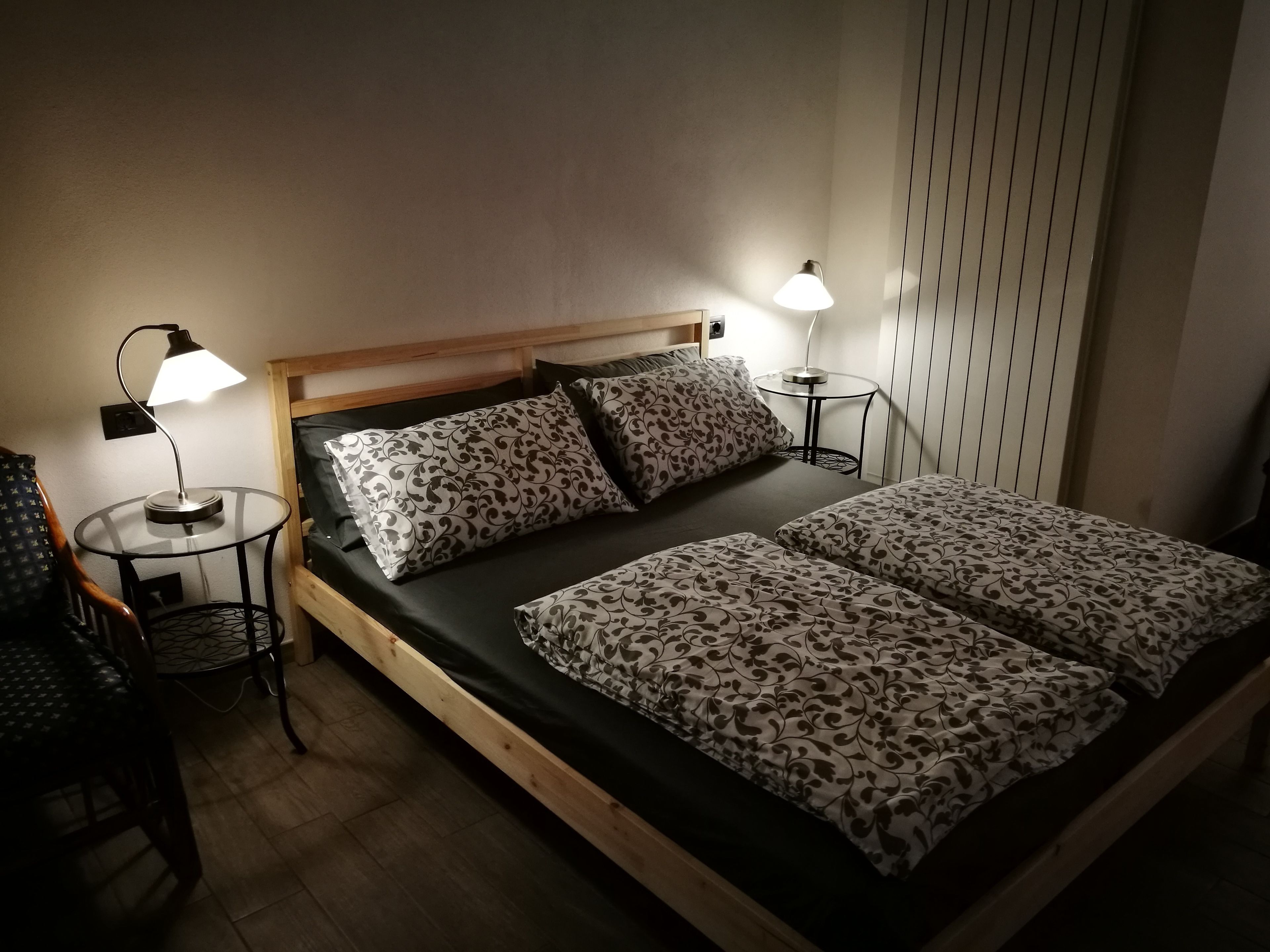 A single bed with a wooden headboard, two bedside lamps, a small round table, and a chair with a metal frame.