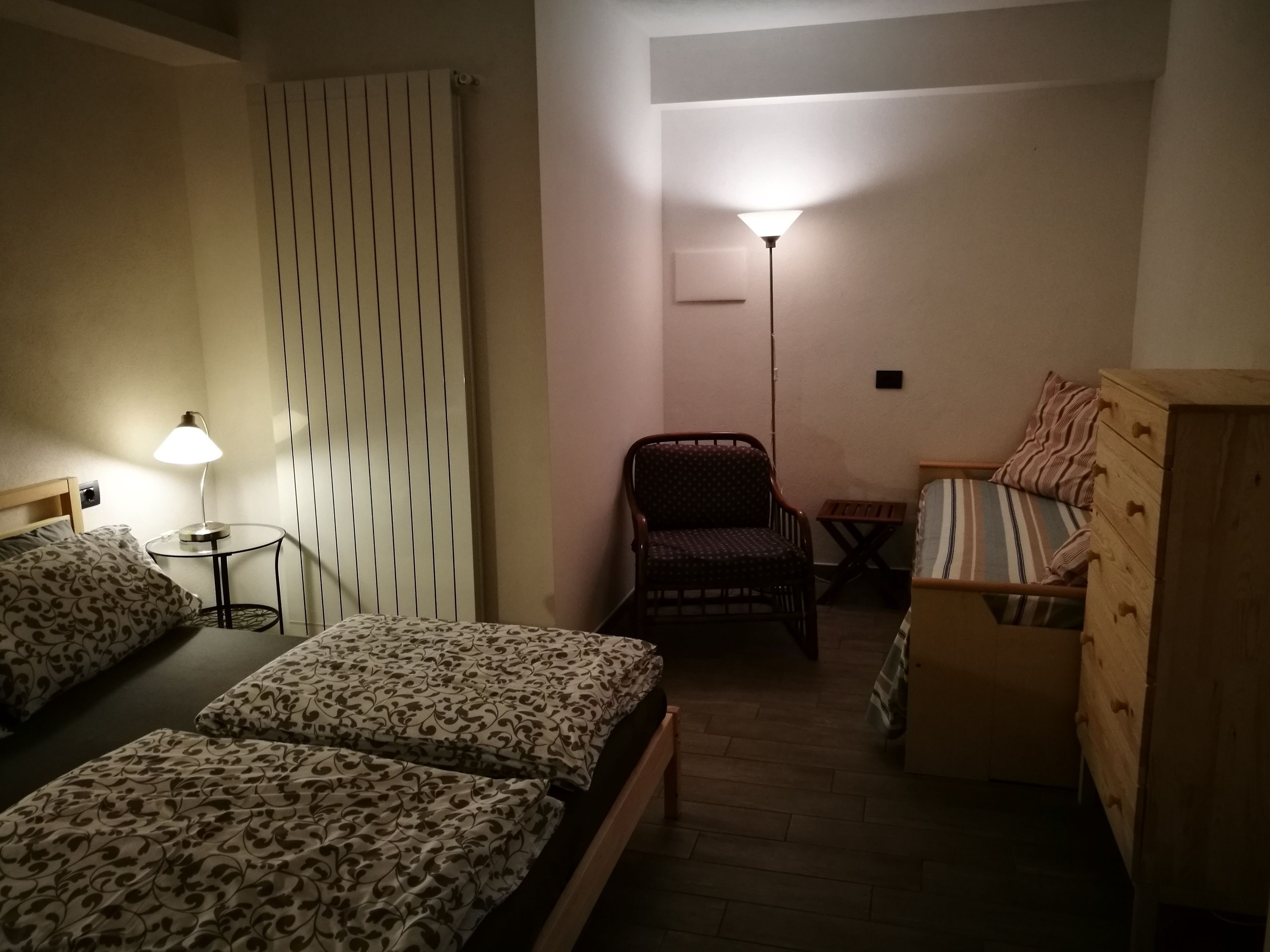 A small, dimly lit room with two beds, a chair, a wooden dresser, and a standing lamp.