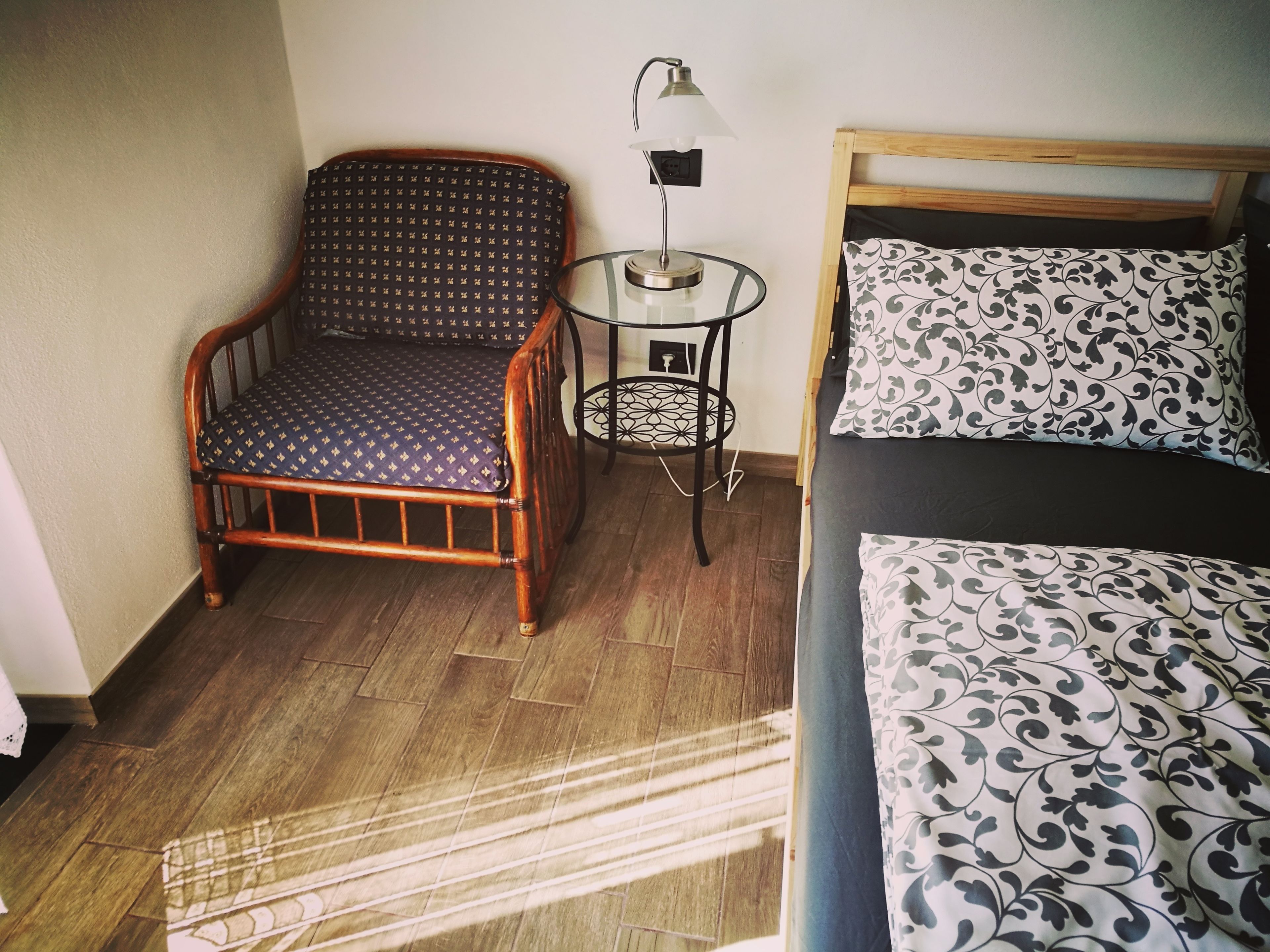 A room with a wooden chair, a small table with a lamp, a bed with patterned bedding, and a wooden floor.