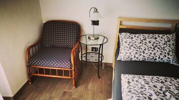 Une pièce avec une chaise en bois, une petite table avec une lampe, un lit avec une literie à motifs et un plancher en bois.