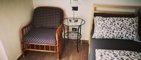 A room with a wooden chair, a small table with a lamp, a bed with patterned bedding, and a wooden floor.