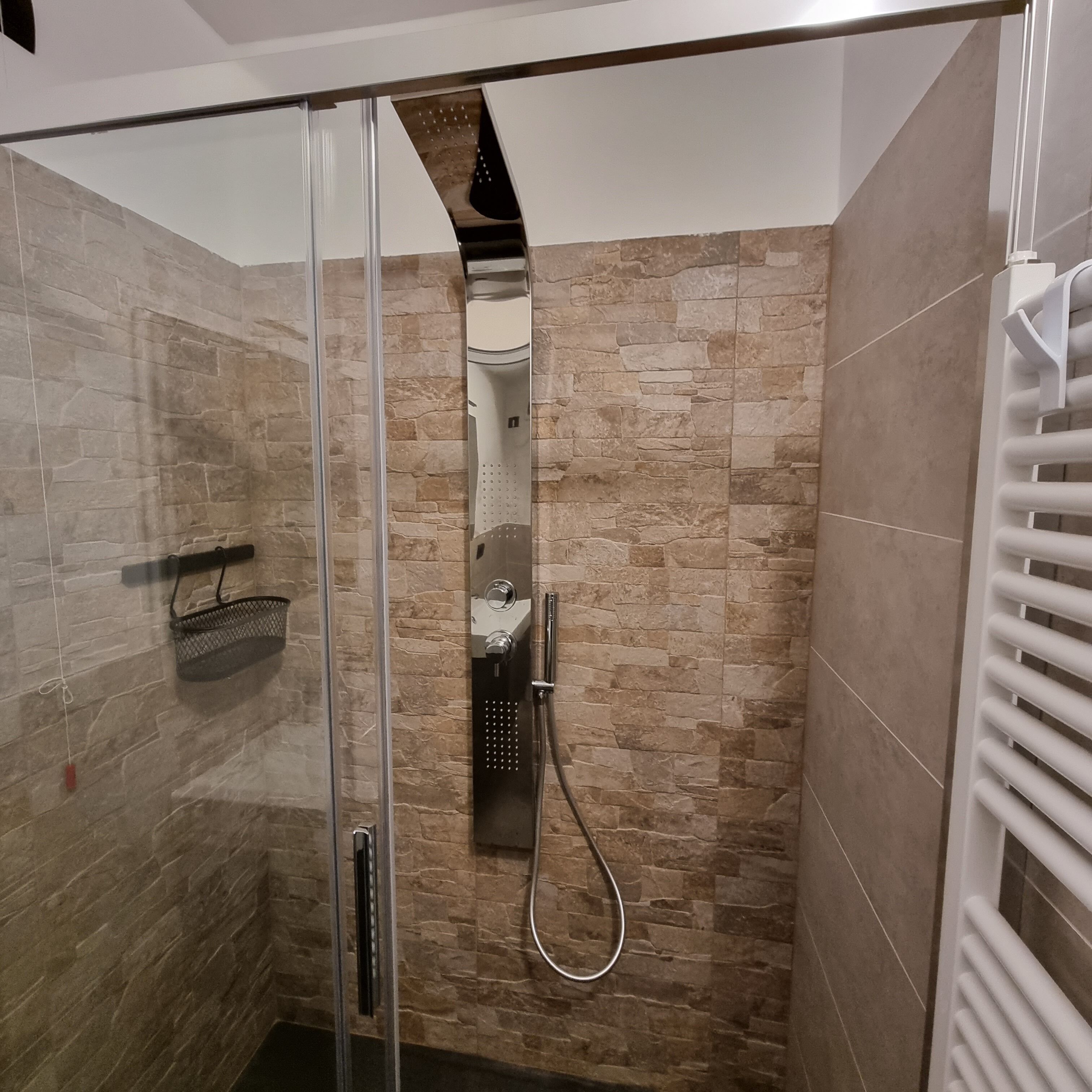 Um chuveiro com divisória de vidro, parede com azulejos de pedra, chuveiro de metal e um porta-toalhas branco.