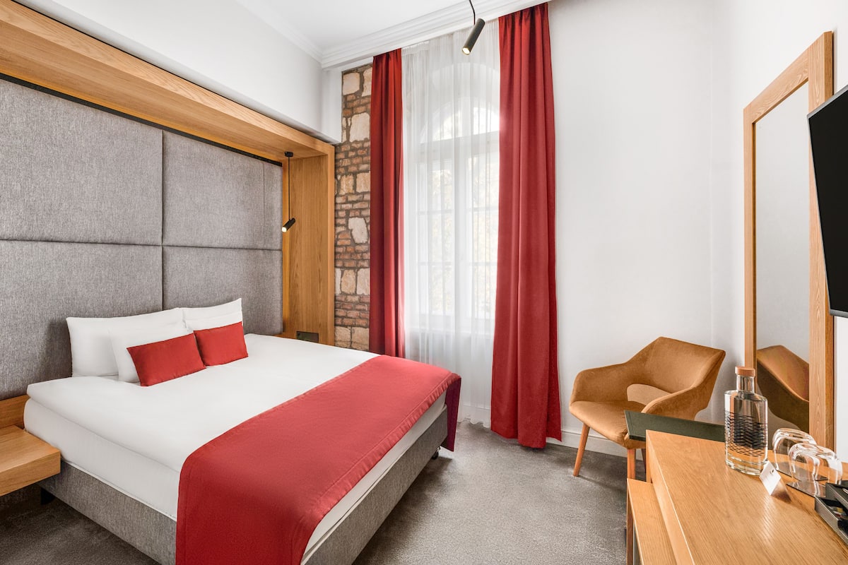 A hotel room with a bed, a wooden nightstand, a chair, a desk with a bottle and glasses, and a large mirror.