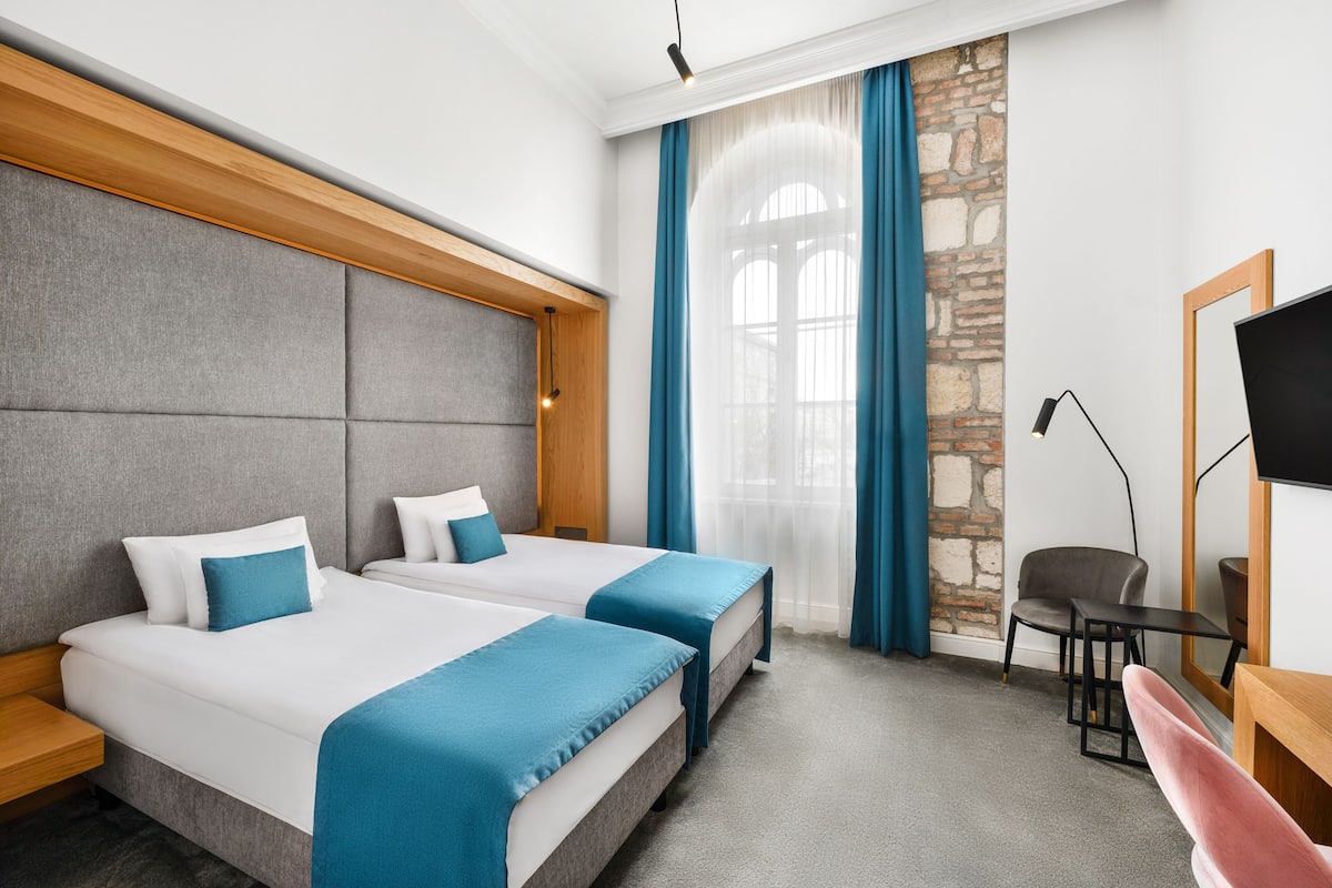 A hotel room with two beds, a stone accent wall, a large window with blue curtains, a desk with a chair, and a TV.