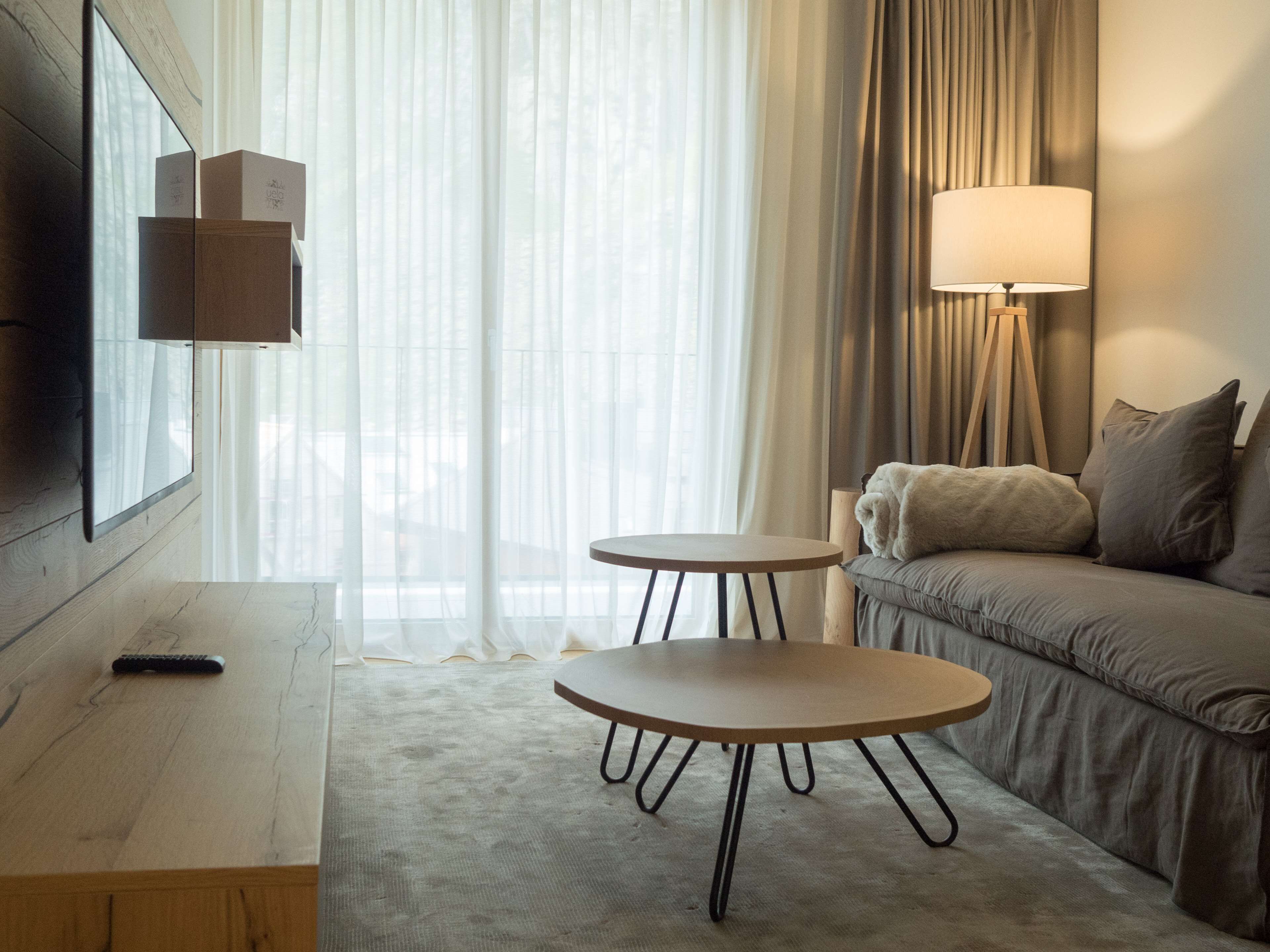 A modern hotel room with a large window, a sofa, two small tables, a remote control, and a lamp.
