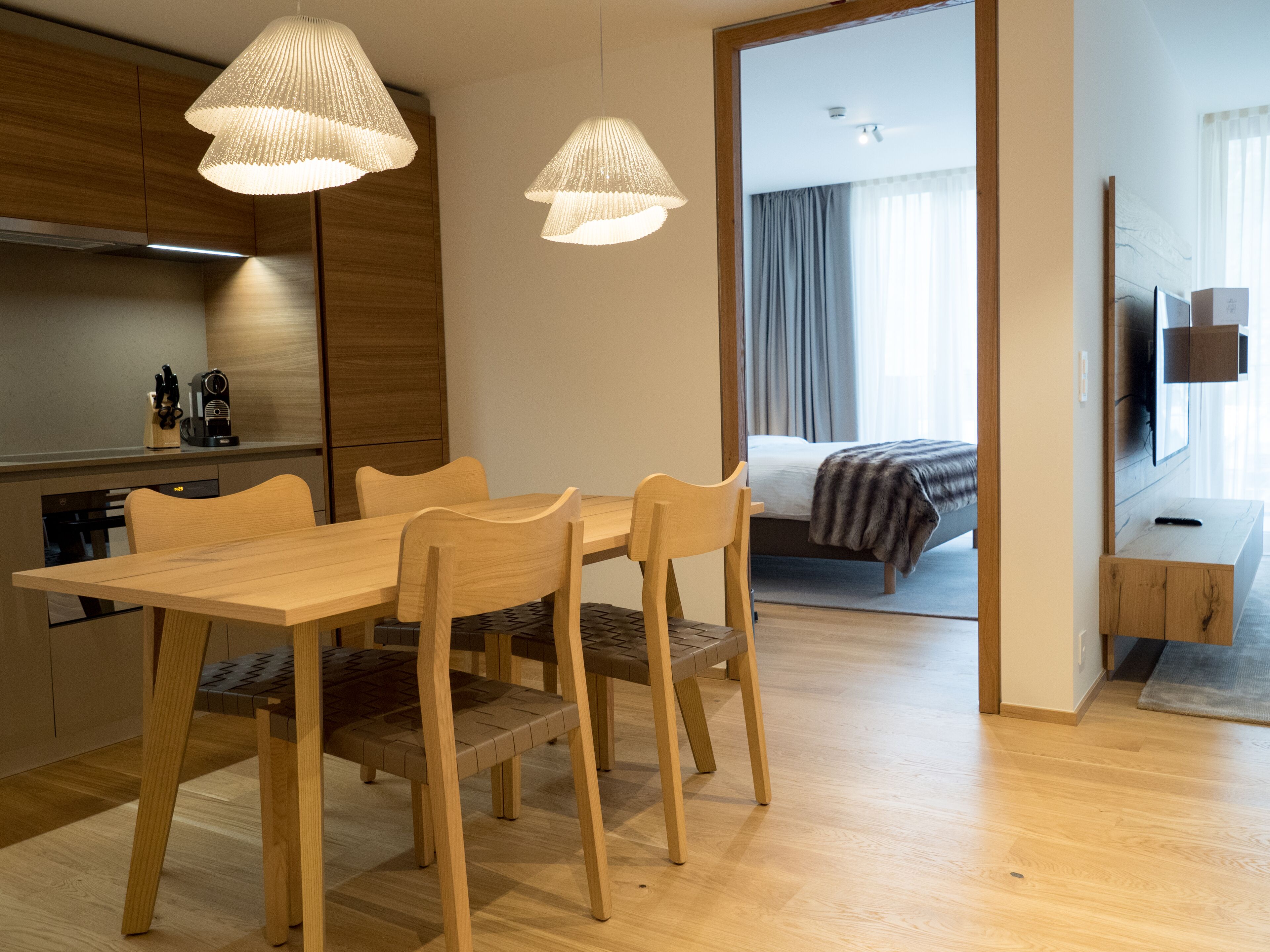 A modern hotel room with a wooden dining table, chairs, and a large bed with a blanket. There are two pendant lights above the table and a flat-screen TV mounted on the wall.