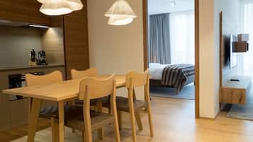 A modern hotel room with a wooden dining table, chairs, and a large bed with a blanket. There are two pendant lights above the table and a flat-screen TV mounted on the wall.