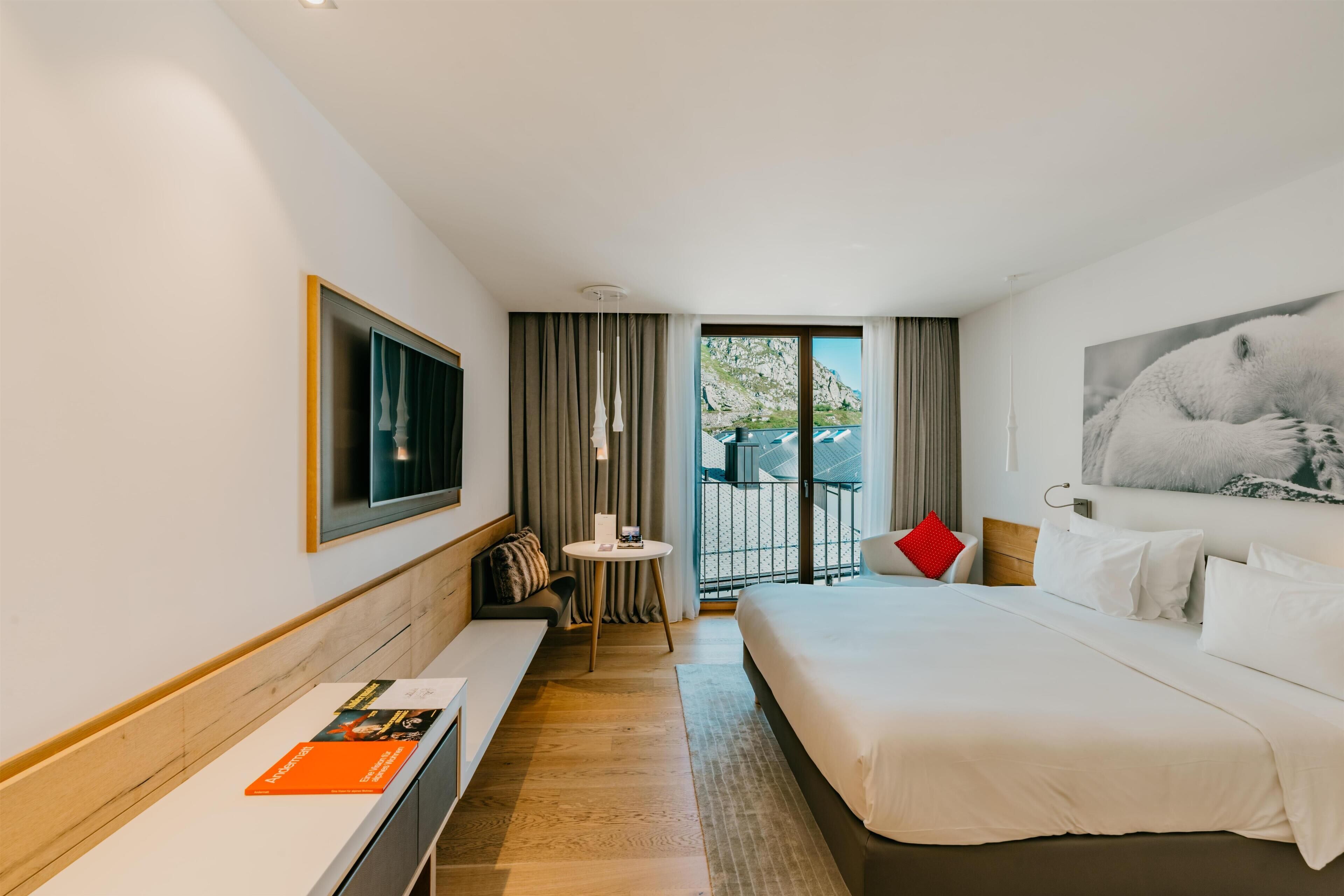 Habitación de hotel moderna con una cama grande, un área de descanso con una mesa y un balcón con vistas a las montañas.