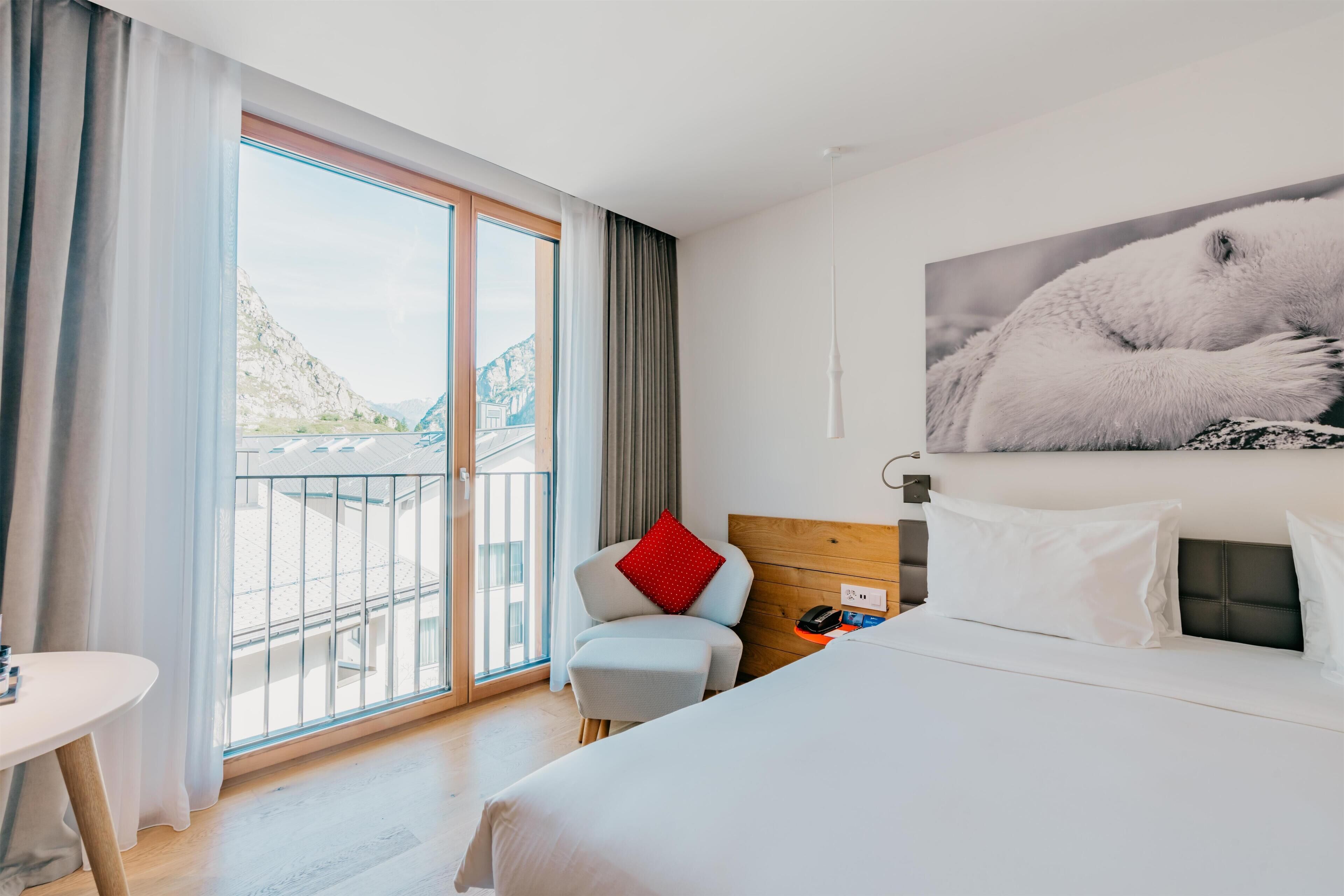 Une chambre d’hôtel comprenant un lit, une chaise, une petite table, une grande fenêtre avec des rideaux et une œuvre d’art accrochée au mur.