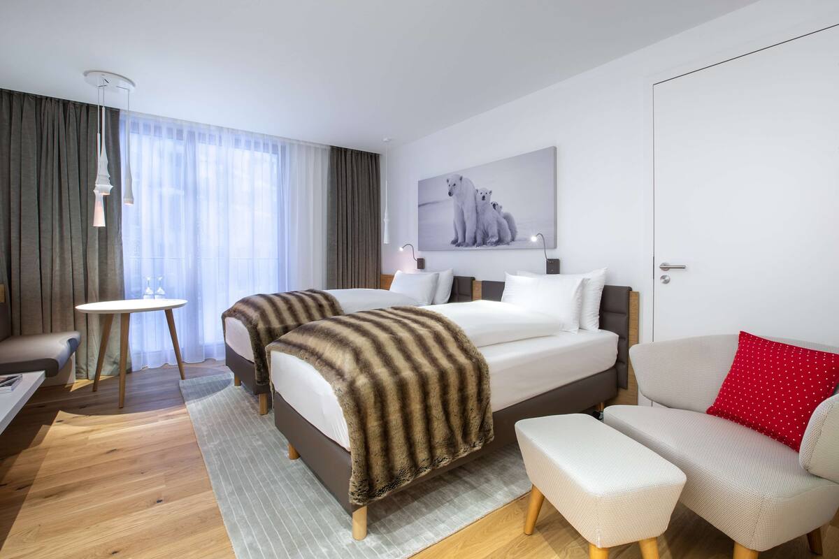 A hotel room with two beds, a sofa, a small table, and a large window with curtains.