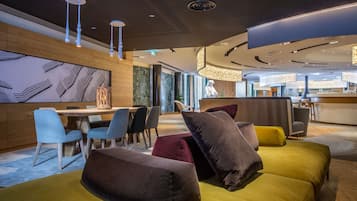 Eine moderne Hotelhalle mit einem Holzbüro, einem Essbereich mit blauen Stühlen und einer Sitzecke mit gelben und braunen Sofas.