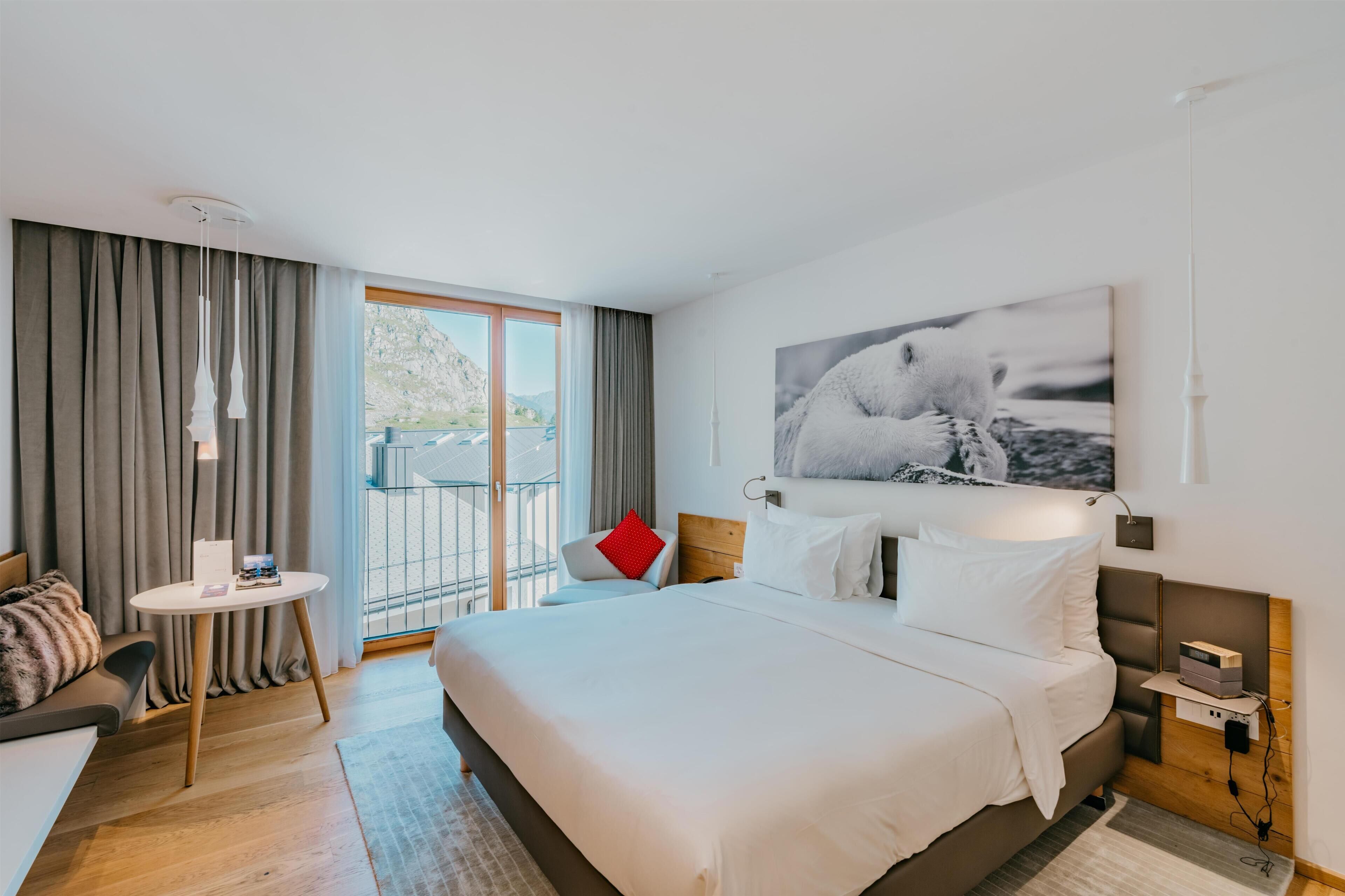 Una habitación de hotel moderna con una cama grande, una mesita redonda, un sofá y un balcón con cortinas.