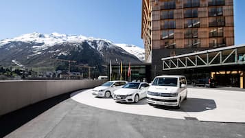A modern building with a curved driveway, parked white cars, and a mountain backdrop.