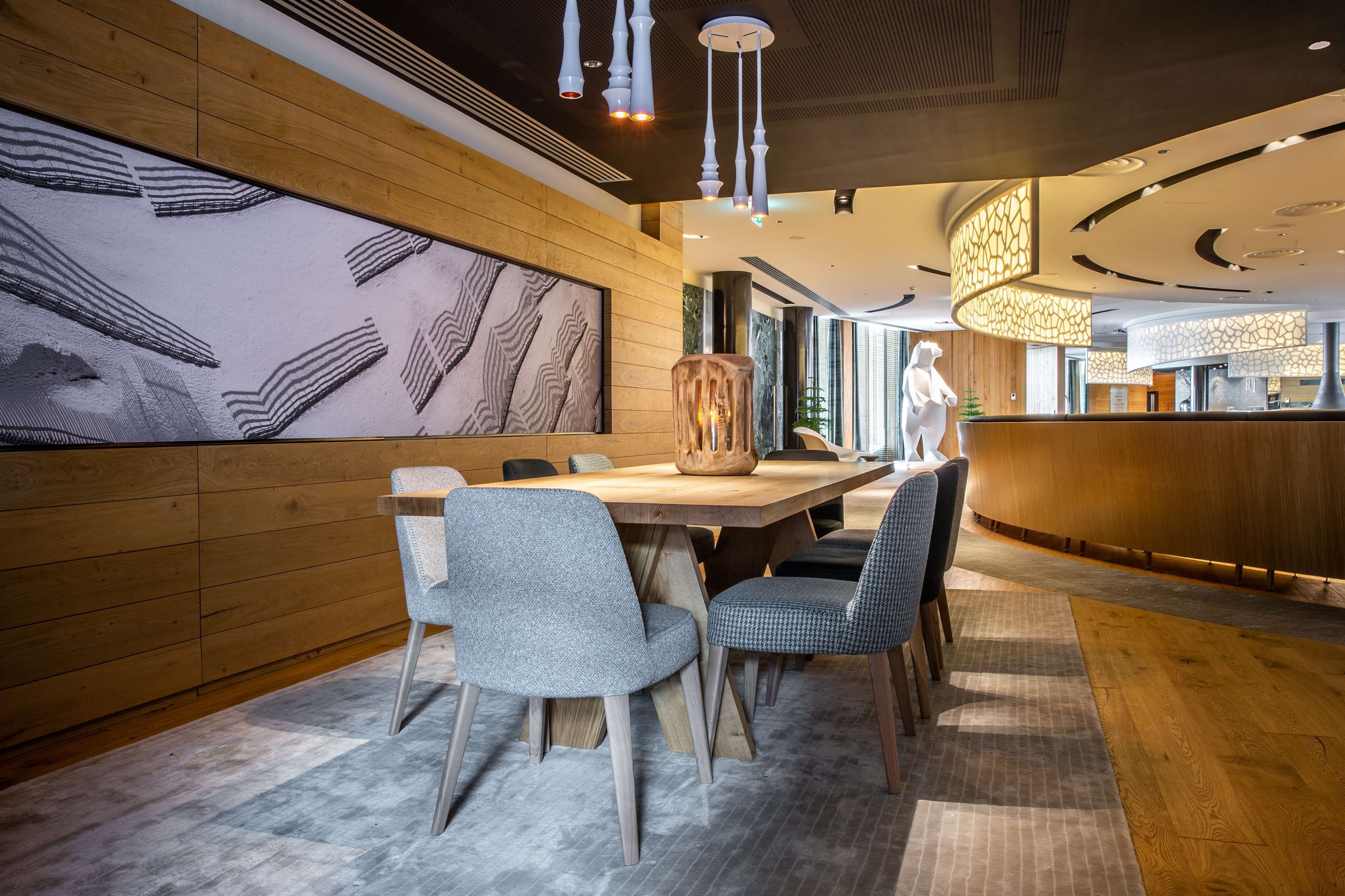 A modern interior with a wooden panel wall, a large abstract artwork, a wooden dining table with chairs, and a curved reception desk.