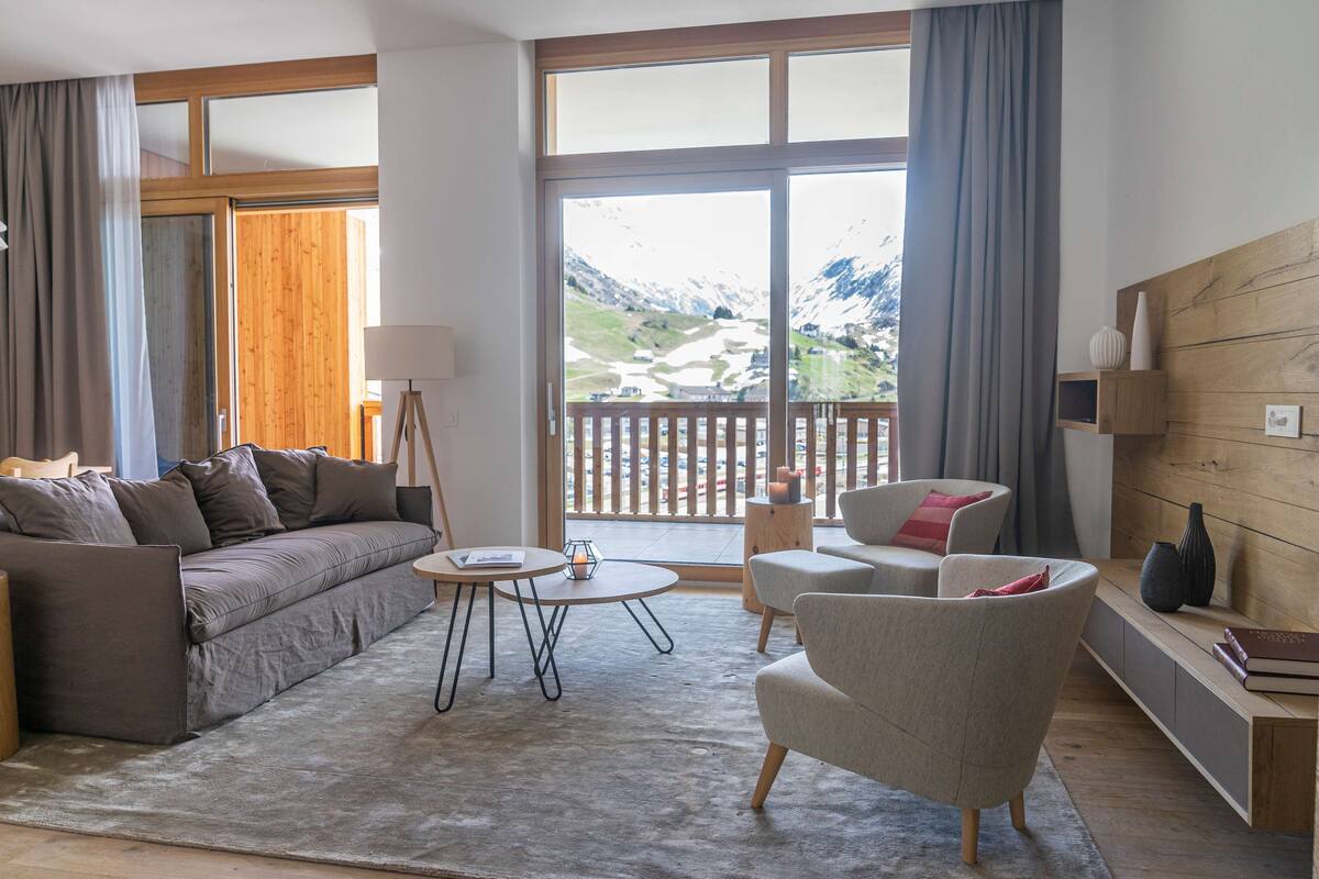 A modern living room with a sofa, armchair, and coffee table, large windows with a mountain view, and a wooden wall panel.