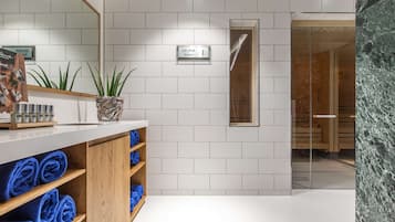 A modern bathroom with white tiles, a wooden vanity with blue towels, a sauna sign, and a glass shower enclosure.