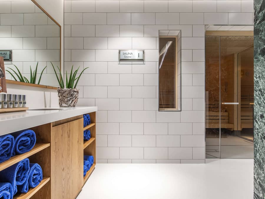 A modern bathroom with white tiles, a wooden vanity with blue towels, a sauna sign, and a glass shower enclosure.
