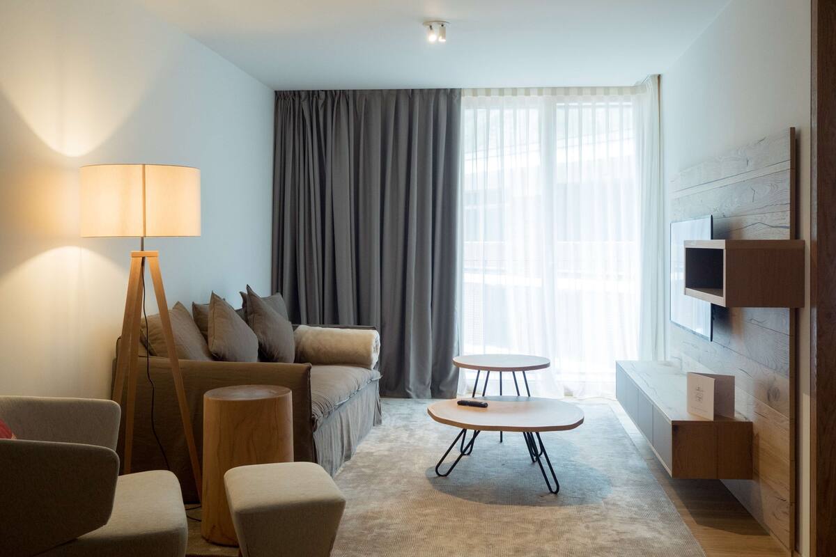 A modern hotel room with a sofa, armchairs, a small table, a floor lamp, and a TV mounted on the wall.