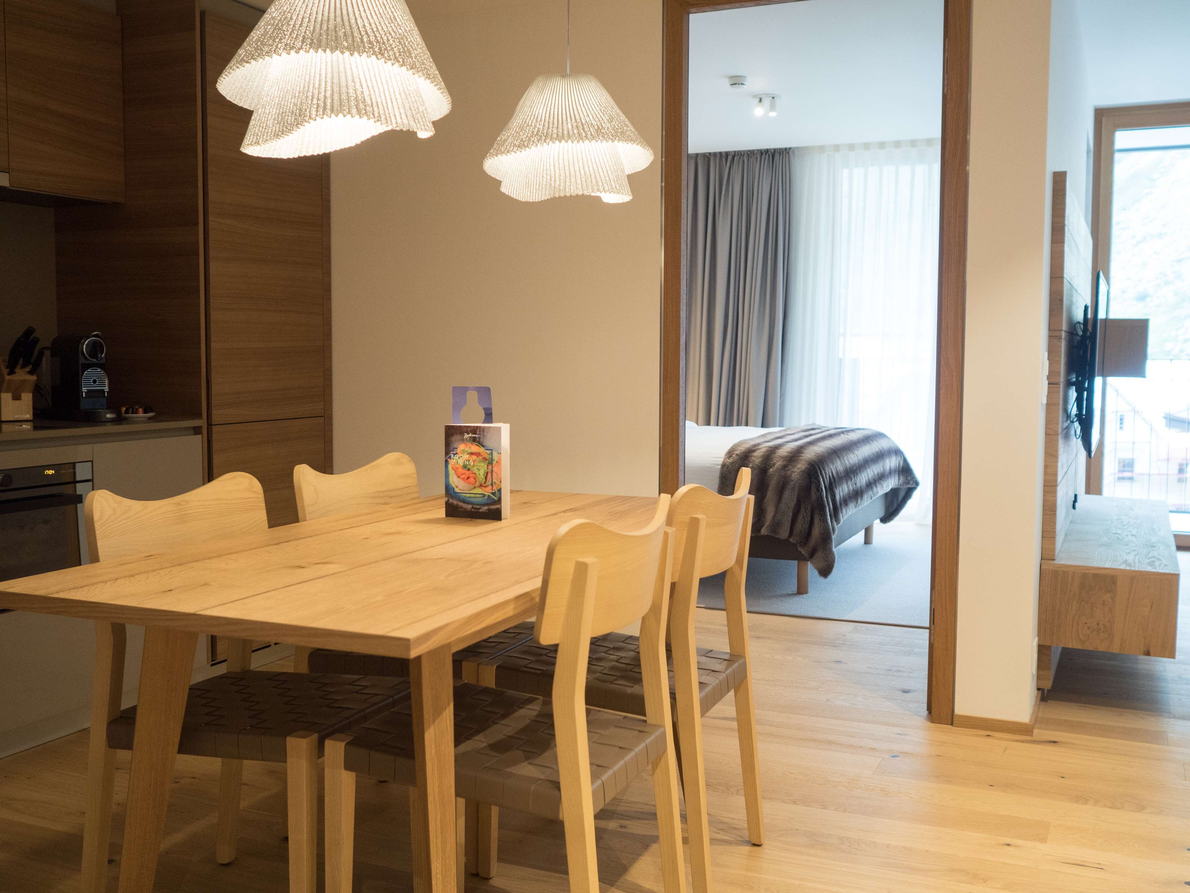 Une chambre d’hôtel moderne avec une table à manger en bois, des chaises et un coin cuisine. On y trouve un lit recouvert d’une couverture grise, et une fenêtre donnant sur l’extérieur.