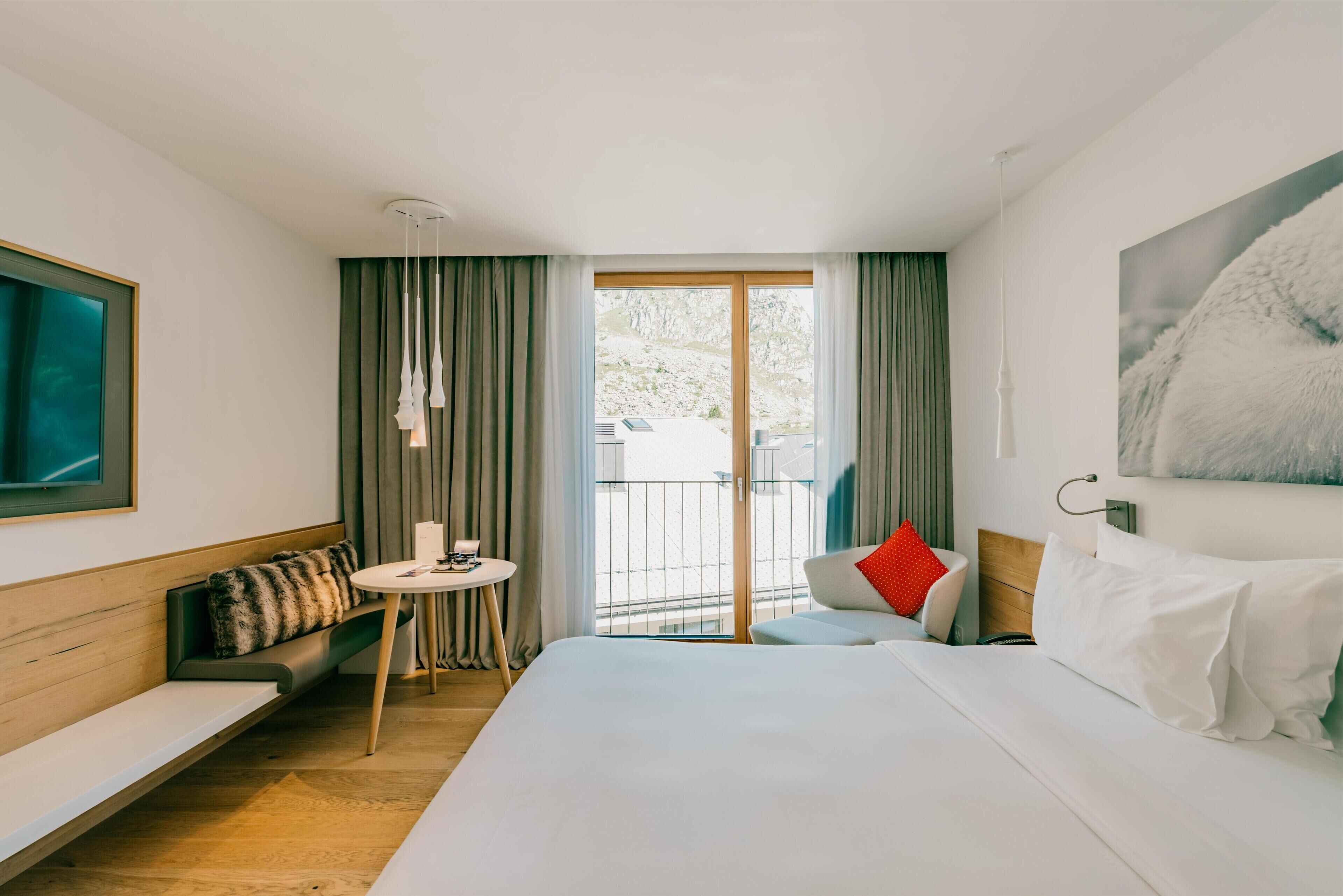 A hotel room with a bed, a sofa, a small table, and a balcony with curtains.