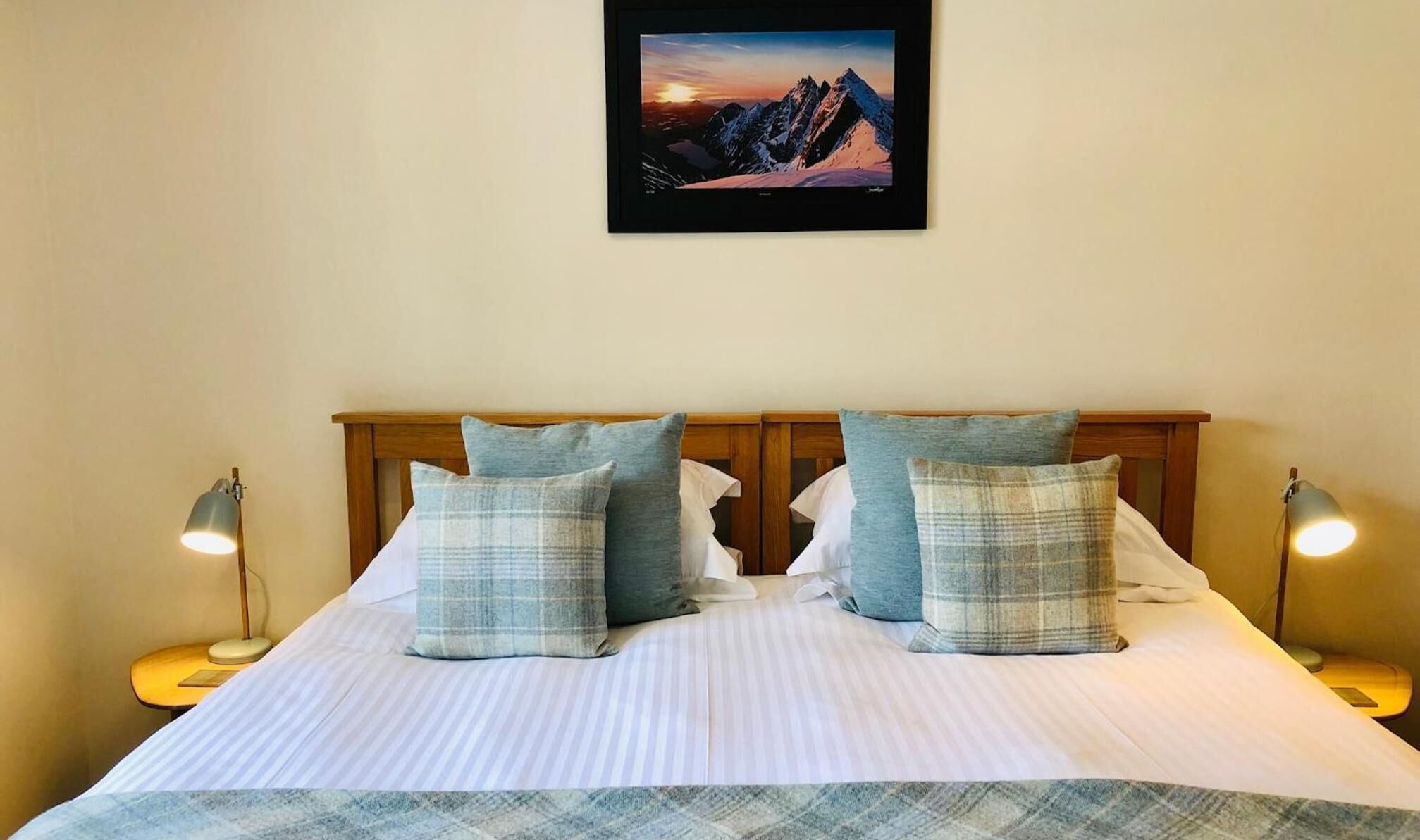 A neatly made bed with a striped bedspread and two pillows, flanked by bedside lamps, in a room with a framed picture of a mountain landscape on the wall.