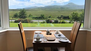 A dining table with chairs, set for two, in a room with a large window offering a view of a green landscape and hills. - Kilmuir Park (Dunvegan)