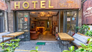 Una entrada de hotel con un letrero que dice "HOTEL" y "Viva La Vita". En el interior, hay mesas y sillas de madera, y un rincón de estar acogedor con cojines.