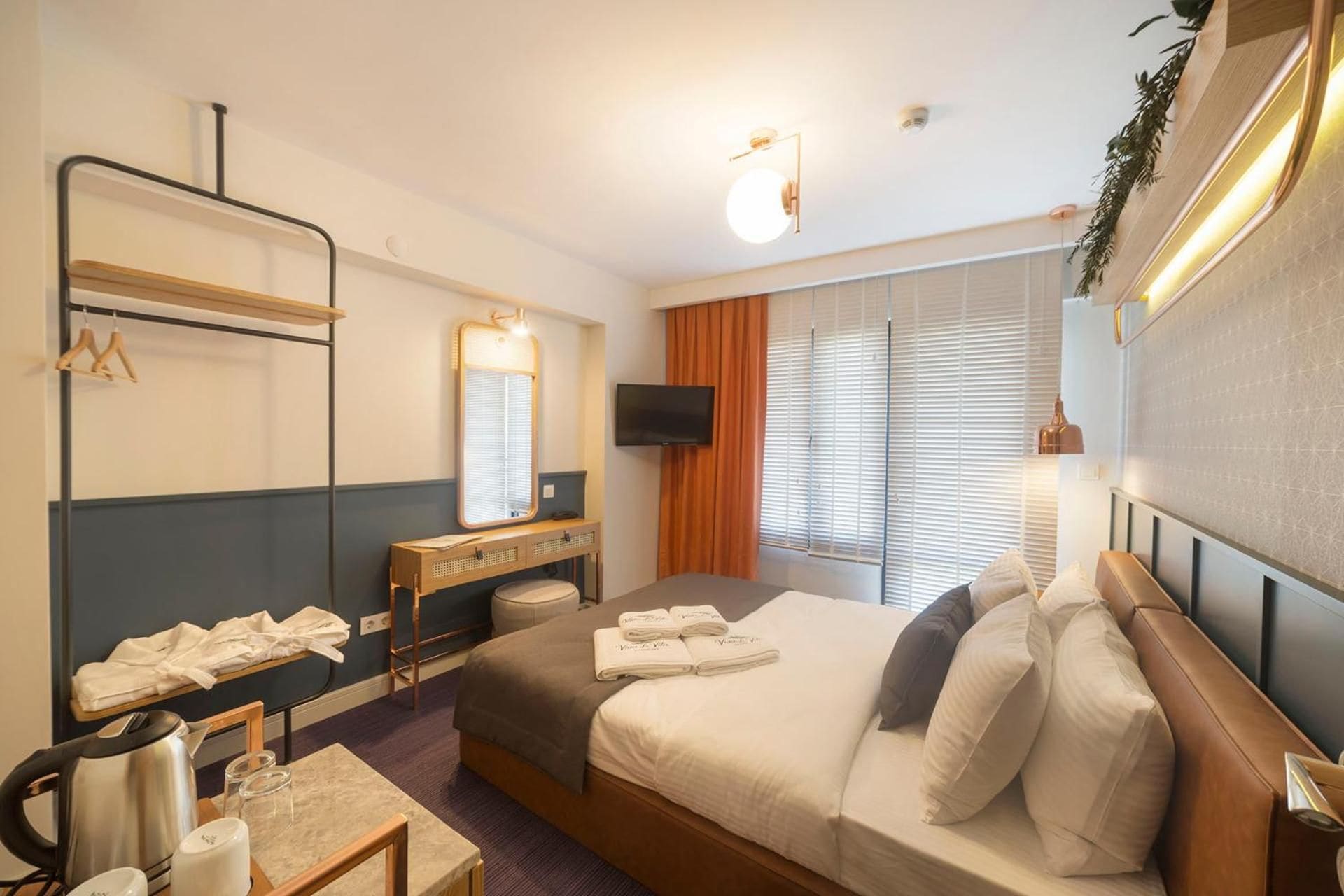 A hotel room with a bed, a television, a mirror, a desk with a kettle, and a window with blinds.