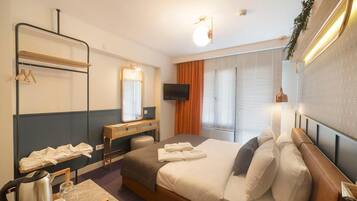Habitación de hotel con cama, televisión, espejo, escritorio con hervidor y ventana con persianas.