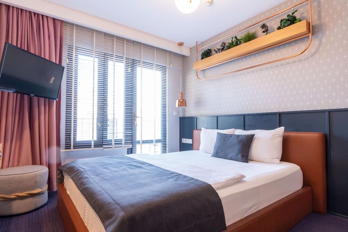 Habitació d'hotel moderna amb un llit gran, televisió penjada, una taula de nit i una finestra amb persianes.