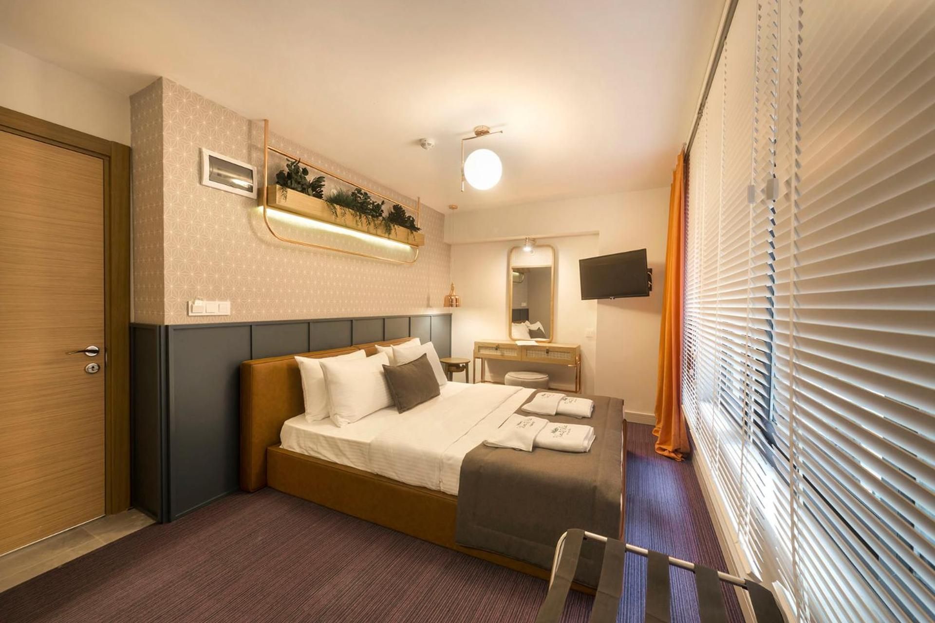 Une chambre d’hôtel équipée d’un lit, d’une table de chevet, d’un miroir, d’une télévision et d’une fenêtre avec des stores.