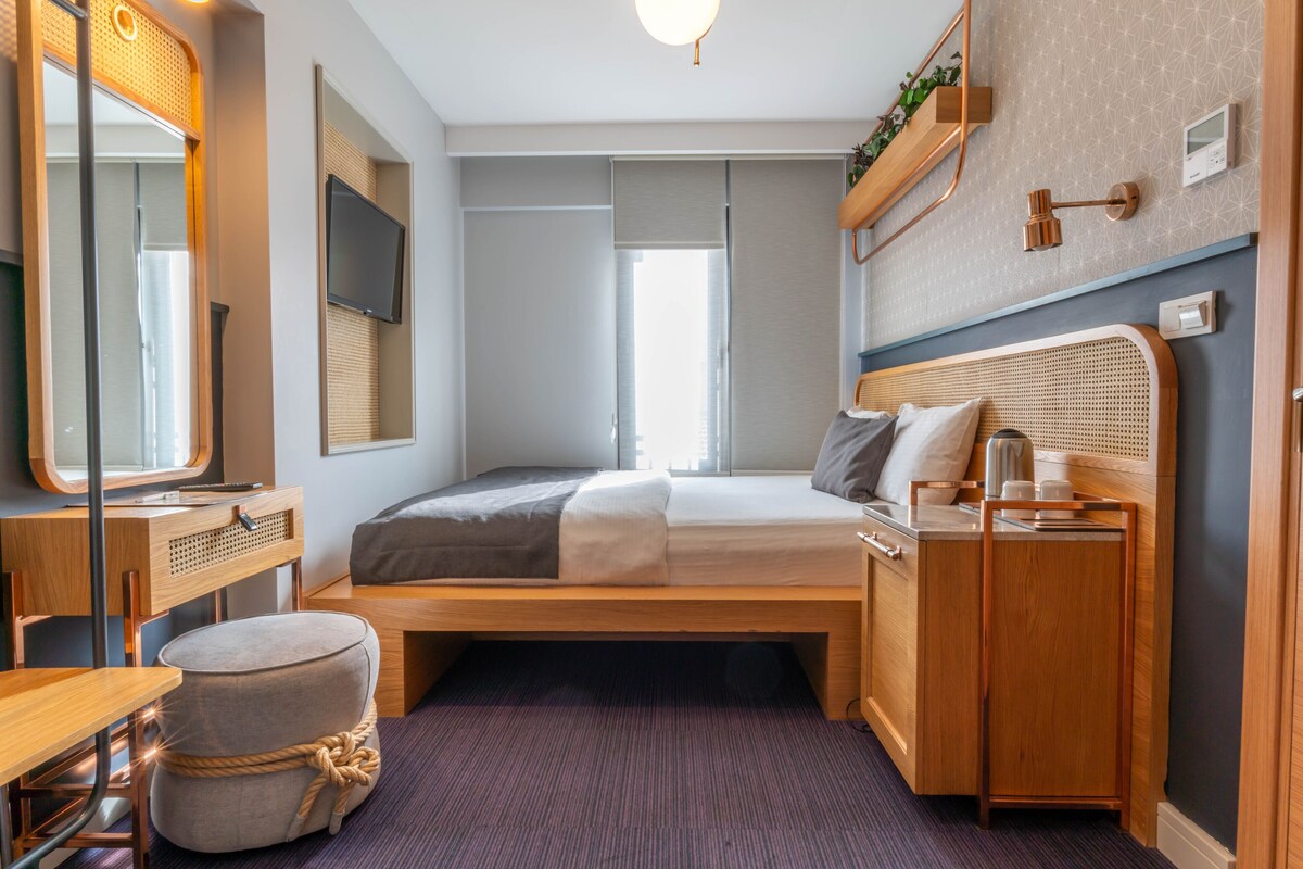 Quarto compacto com cama, escrivaninha, cadeira, um banquinho pequeno, televisão e janela.