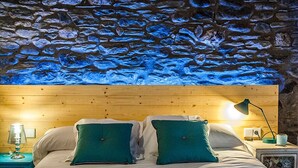 A bedroom with a stone wall, wooden headboard, two green pillows, a white bedspread, a nightstand with a lamp, and a framed picture. - Cal Batlle Casa Rural (La Cortinada)