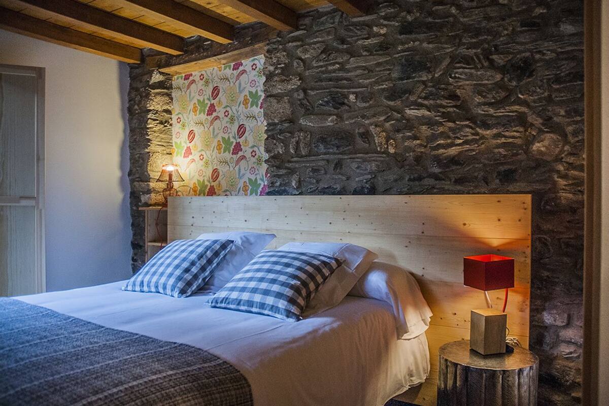 A bedroom with a stone wall, a wooden headboard, a bed with checkered pillows, and a red lamp on a wooden stool.