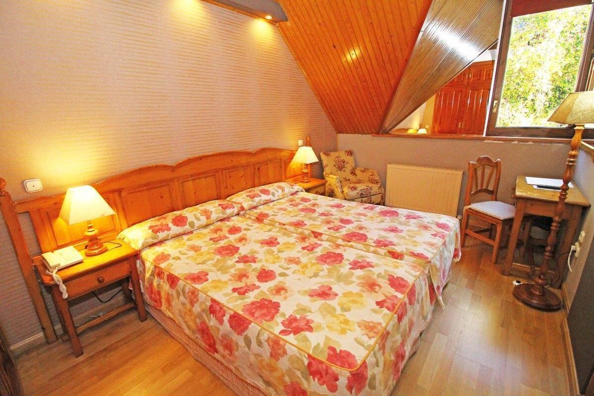 Quarto com teto de madeira, cama com roupa de cama floral, duas mesas de cabeceira com abajures, uma cadeira e uma escrivaninha com luminária.