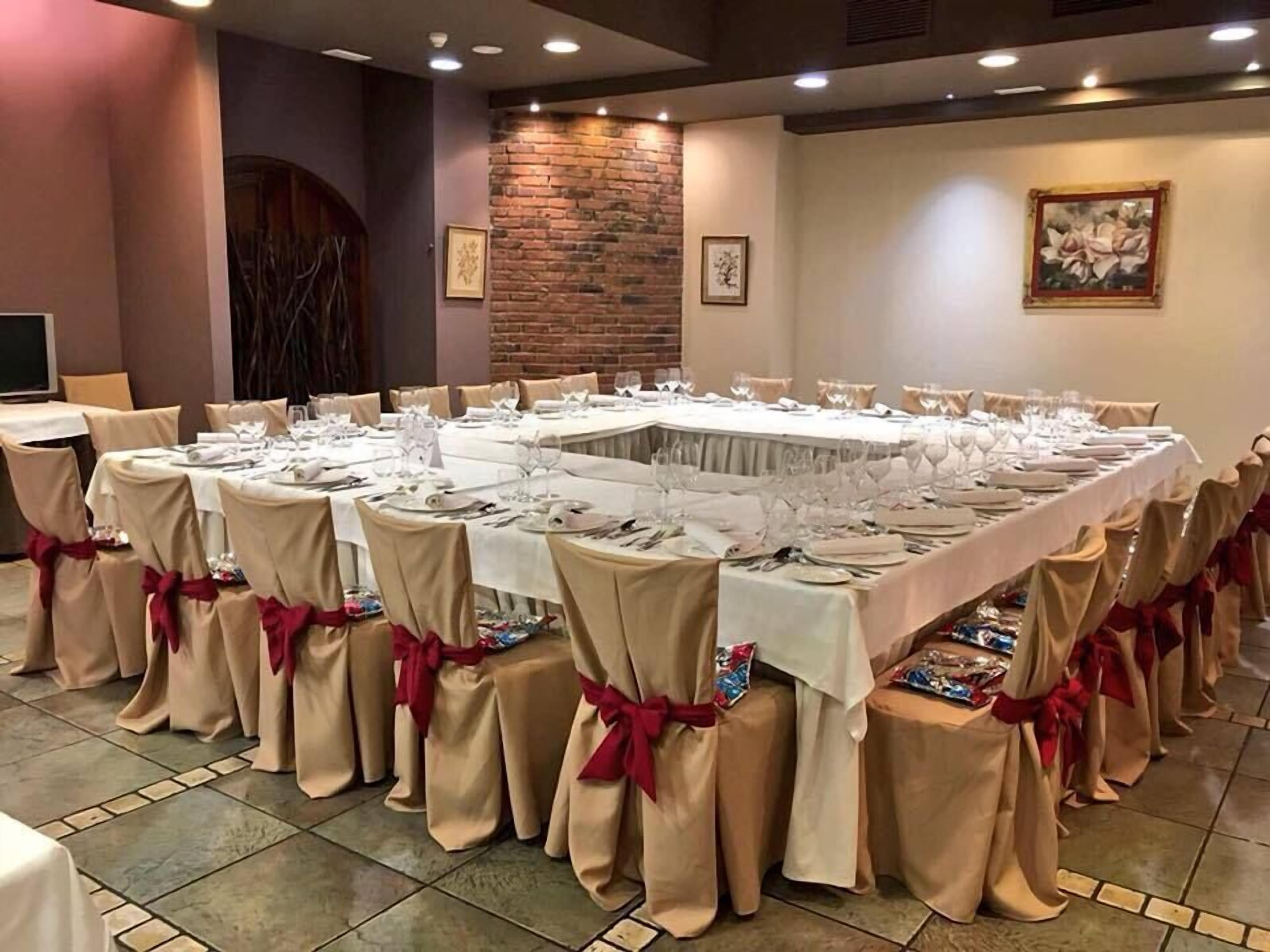 A banquet hall with a long rectangular table set for a formal event, surrounded by chairs with beige covers and red sashes.