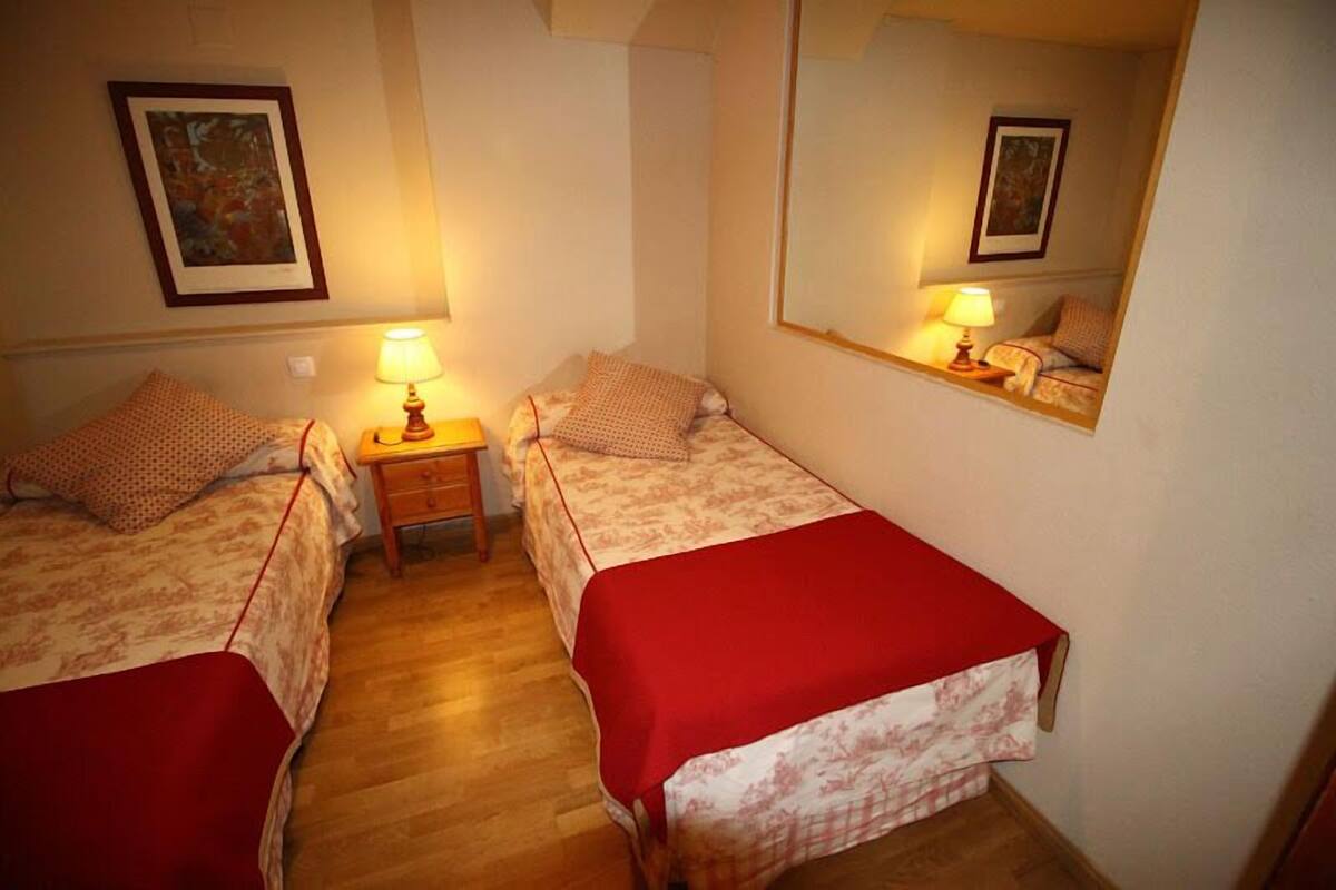 Um quarto pequeno e acolhedor com duas camas de solteiro, cada uma com um cobertor vermelho, uma mesa de cabeceira com abajur e um espelho grande na parede.