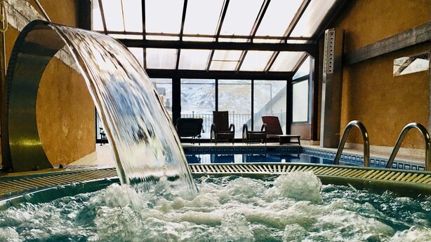 A hot tub with a waterfall feature, surrounded by lounge chairs and a view of a snowy landscape through large windows. - Villa De Sallent 3 (Sallent de Gallego)