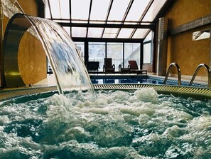 Un jacuzzi con caída de agua, rodeado de sillas de descanso y vistas a un paisaje nevado a través de amplios ventanales.