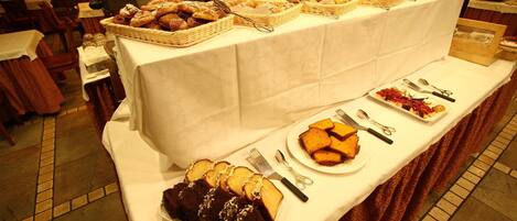 Una mesa de buffet con diversos productos horneados y postres, incluyendo pan, pasteles y repostería, instalada en una sala con paredes de ladrillo y suelo de baldosas.