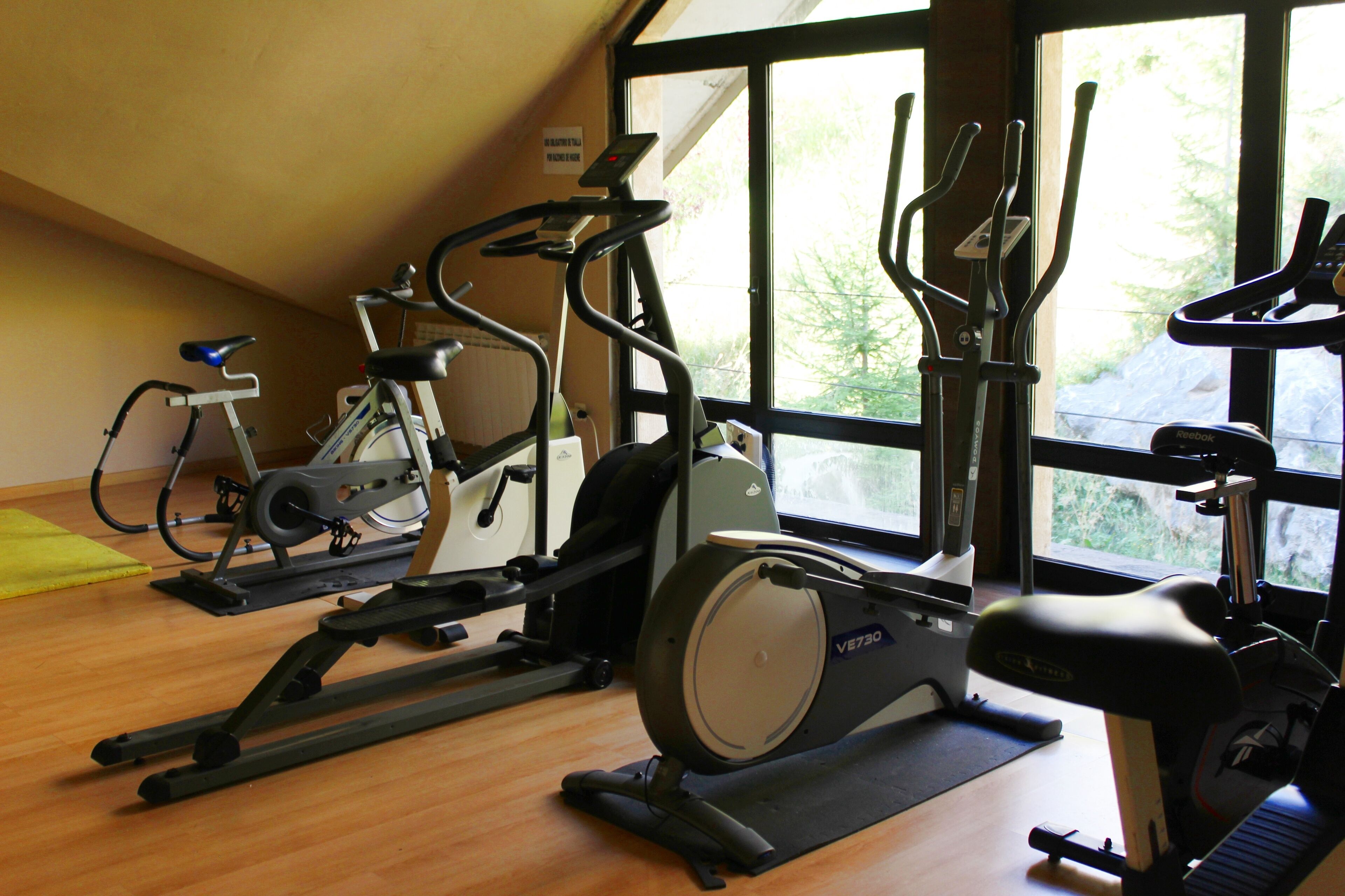 Un gimnasio con diversos equipos de ejercicio, como bicicletas estáticas y elípticas, con amplias ventanas que proporcionan luz natural.
