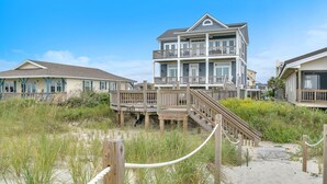 Une propriété située en bord de mer, avec une maison à plusieurs niveaux dotée d’une terrasse en bois et d’un escalier menant à un balcon.