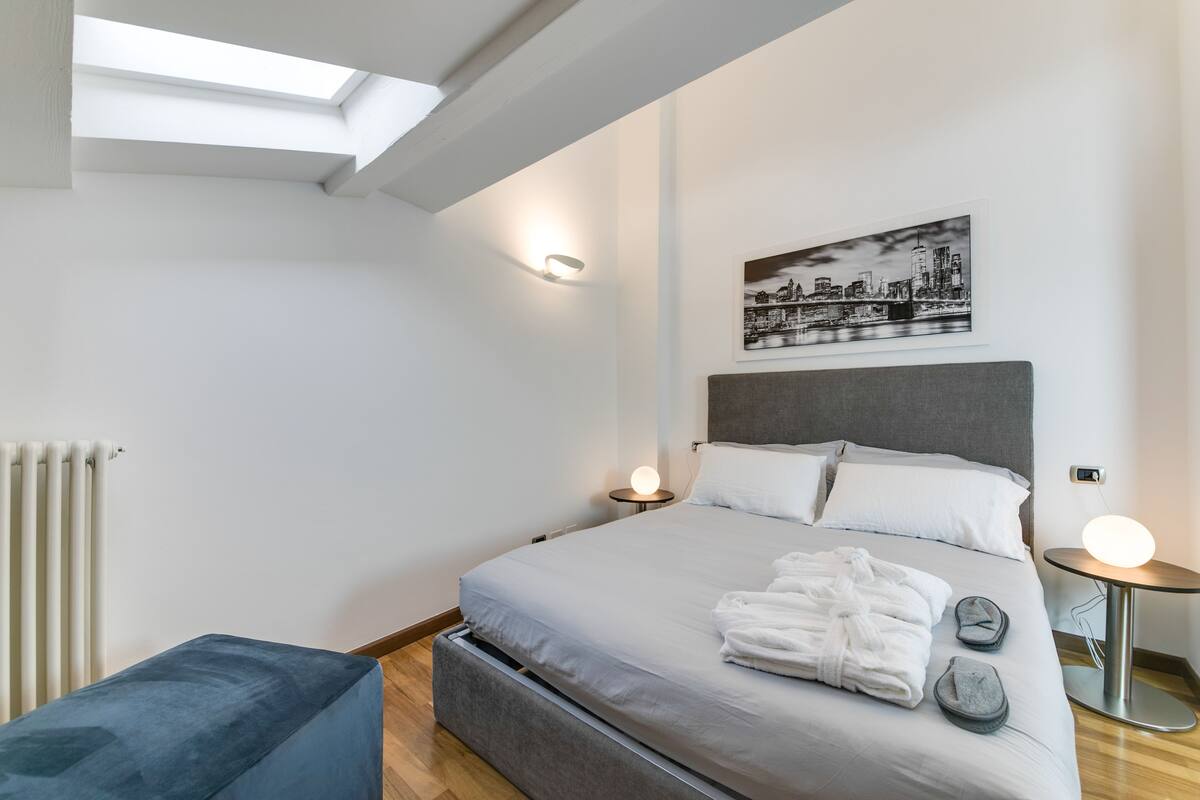 A modern bedroom with a skylight, a bed with white linens, a cityscape artwork, and a bedside table with a lamp.