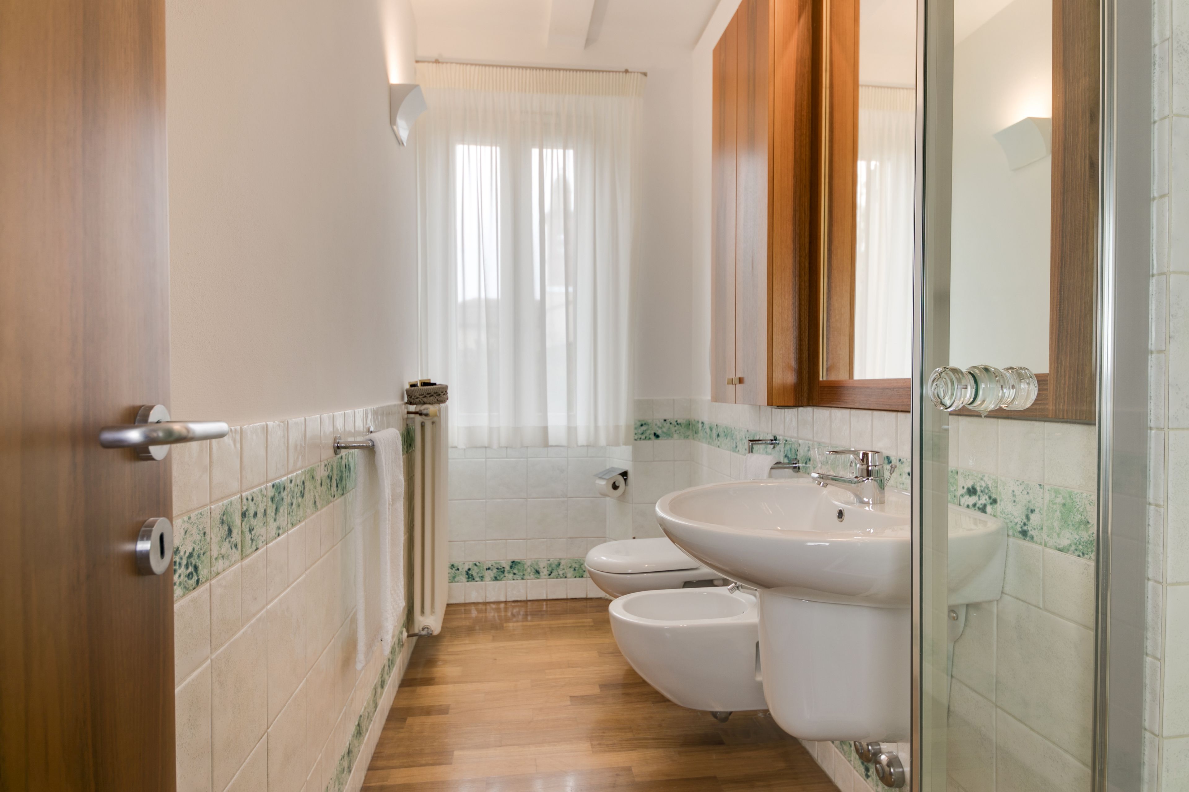 A bathroom with a wooden floor, white tiles, a glass shower enclosure, a double sink, and a toilet.