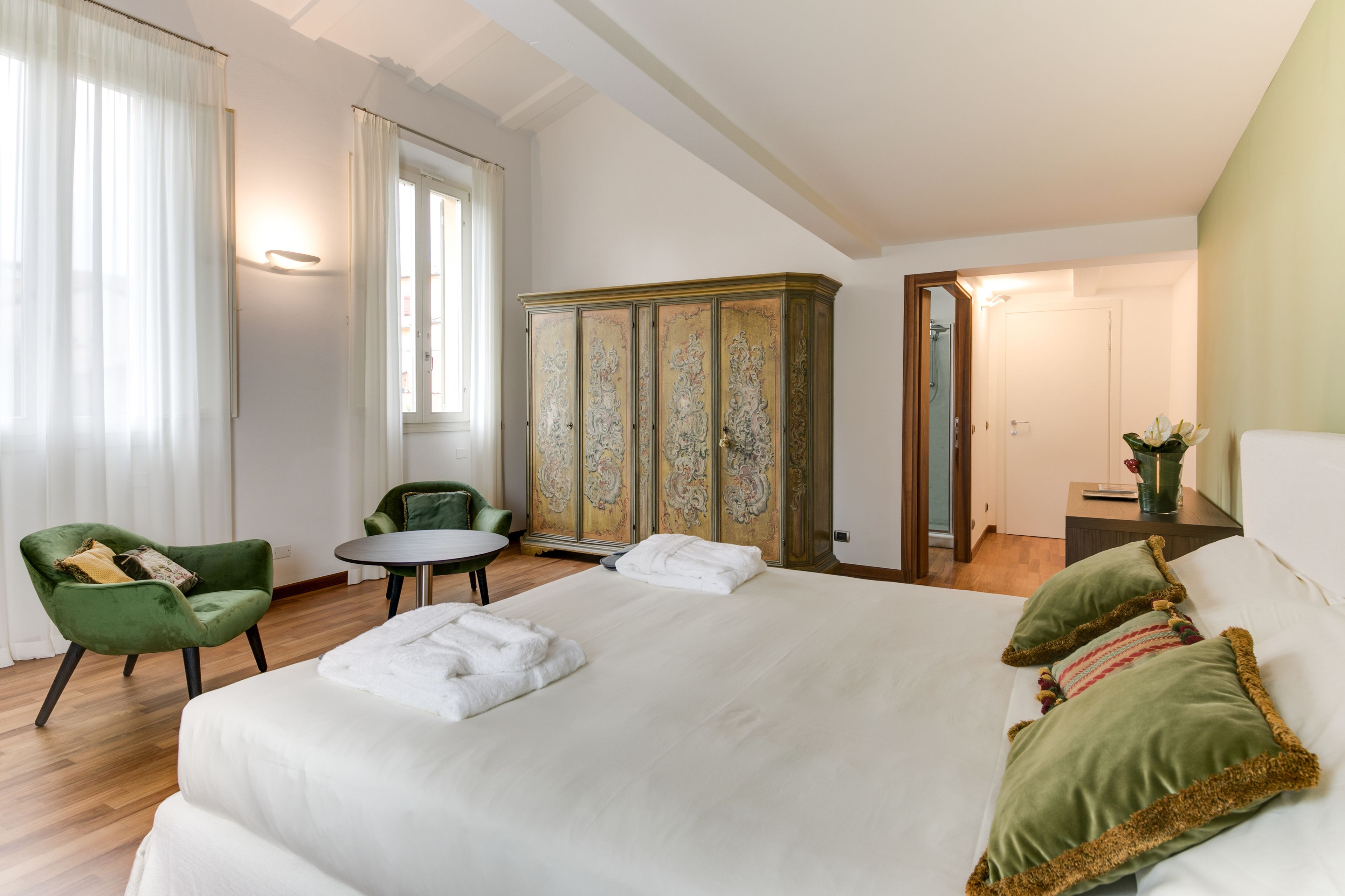 A hotel room with a large bed, a green armchair, a round table, and a wardrobe with ornate doors.