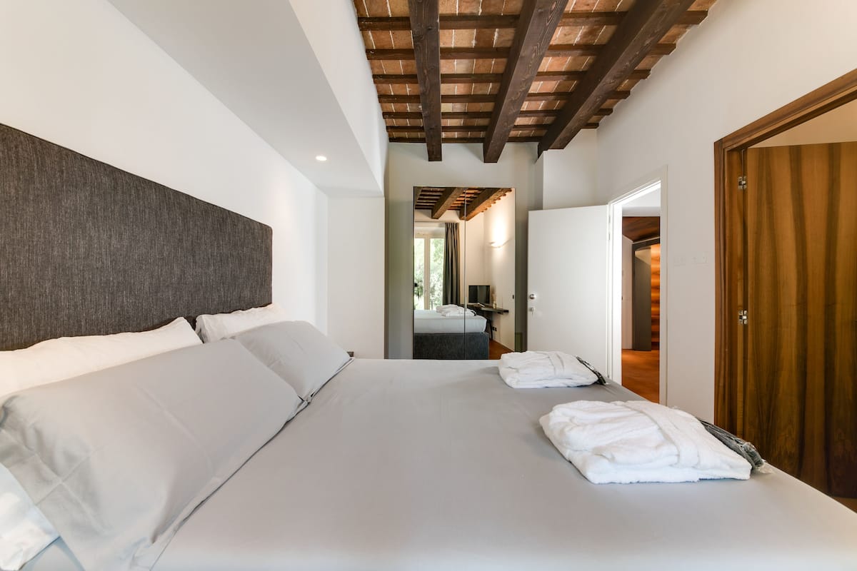 A modern hotel room with a large bed, wooden ceiling beams, a wooden door, and a view into the bathroom.