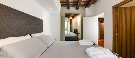 A modern hotel room with a large bed, wooden ceiling beams, a wooden door, and a view into the bathroom.