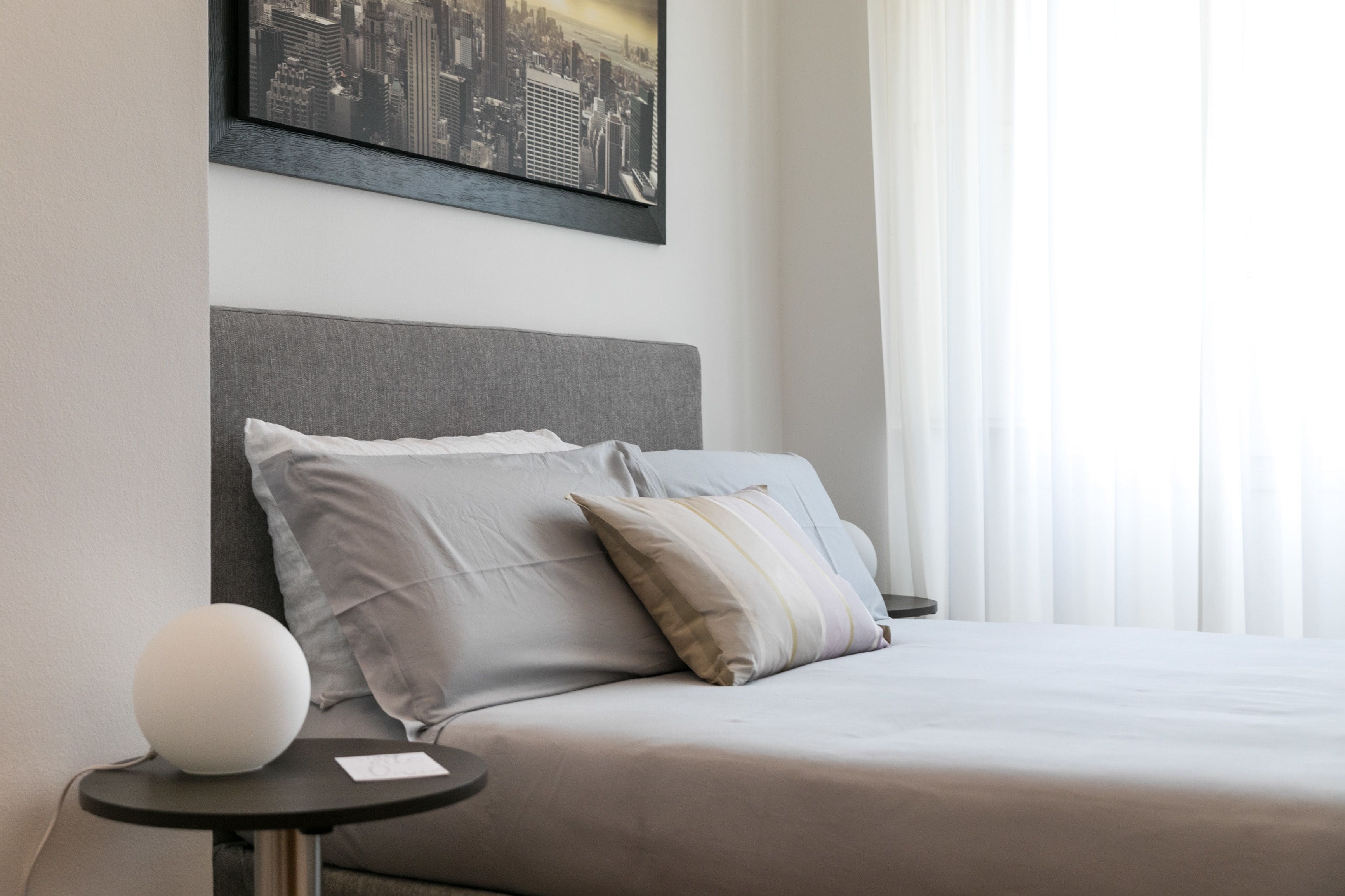 A neatly made bed with a grey headboard, a bedside table with a white spherical object, and a framed cityscape picture on the wall.