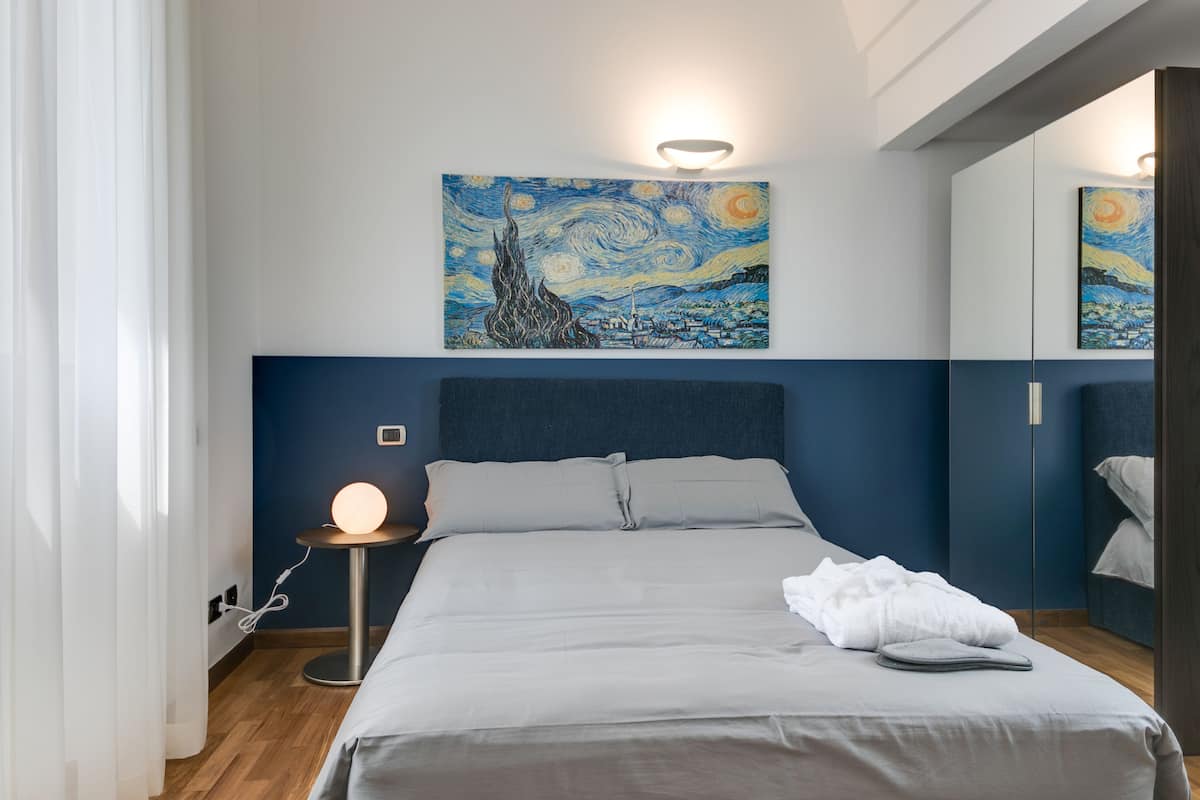 A neatly made bed with a white comforter and pillows, a bedside table with a lamp, and a wall painting above the bed.